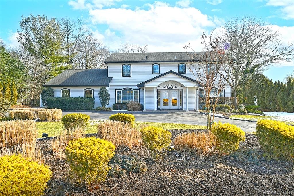 Large white two-story house with black roof, surrounded by shrubs and trees, with a curved driveway leading to the front entrance, in a landscaped yard with yard lighting.
