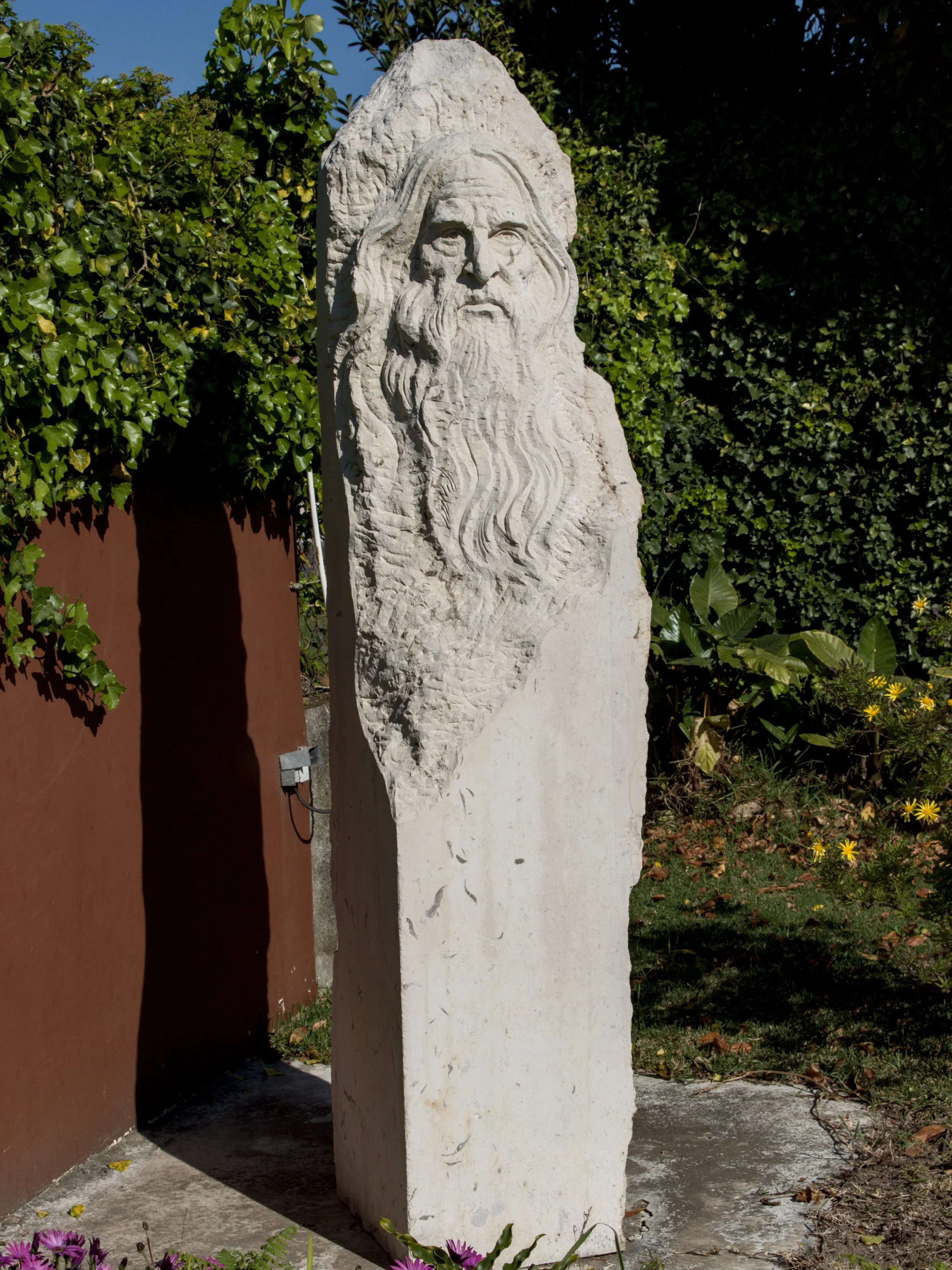 Escultura de pedra com cabelos longos e barba, localizada ao ar livre entre plantas verdes.