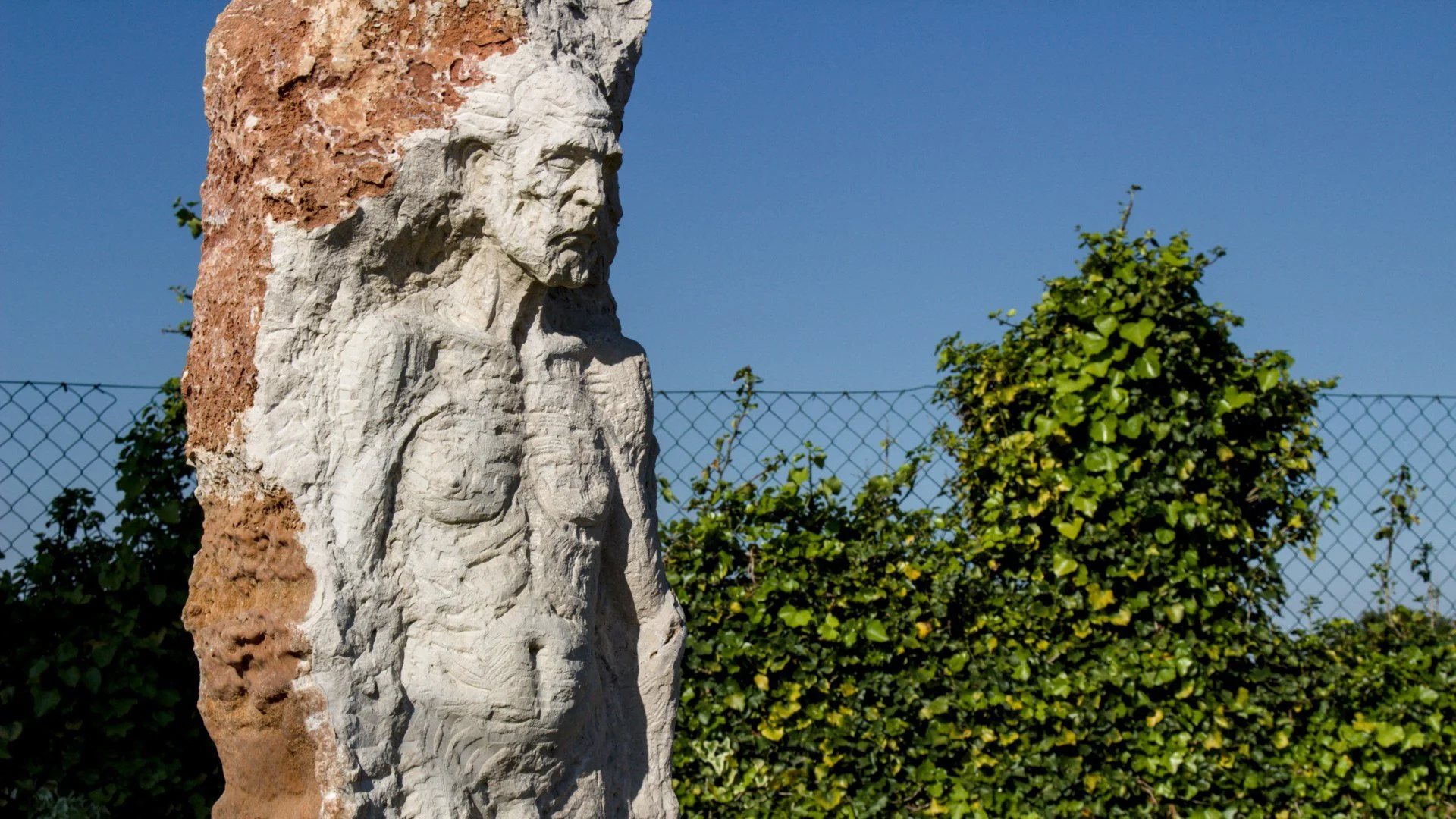 Estátua de um homem com barba e expressão pensativa, esculpida em pedra, ao ar livre com céu azul, arbustos verdes.