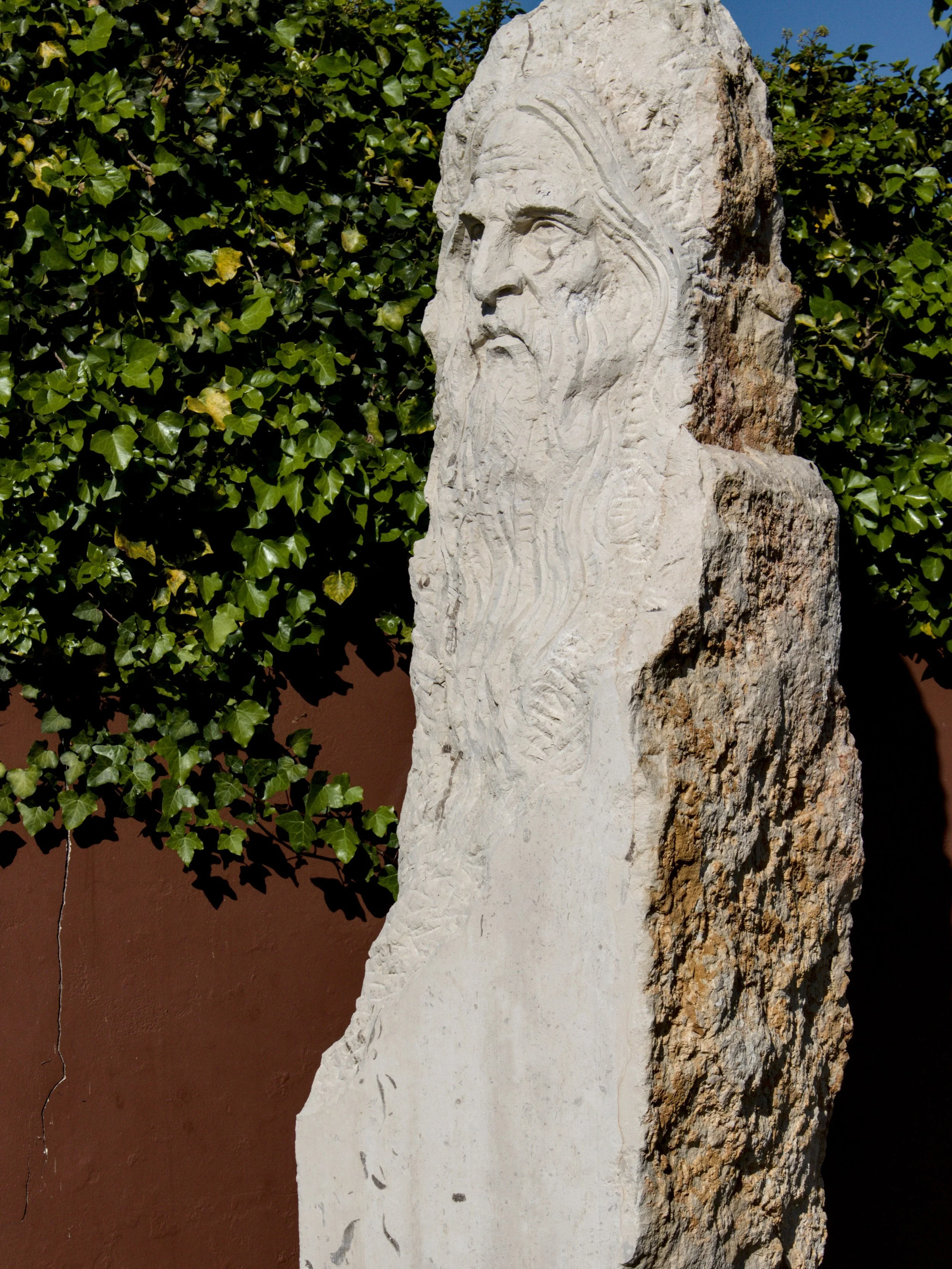 Estátua de pedra de um homem com barba longa e cabeça de perfil, contra um fundo de folhagem verde.
