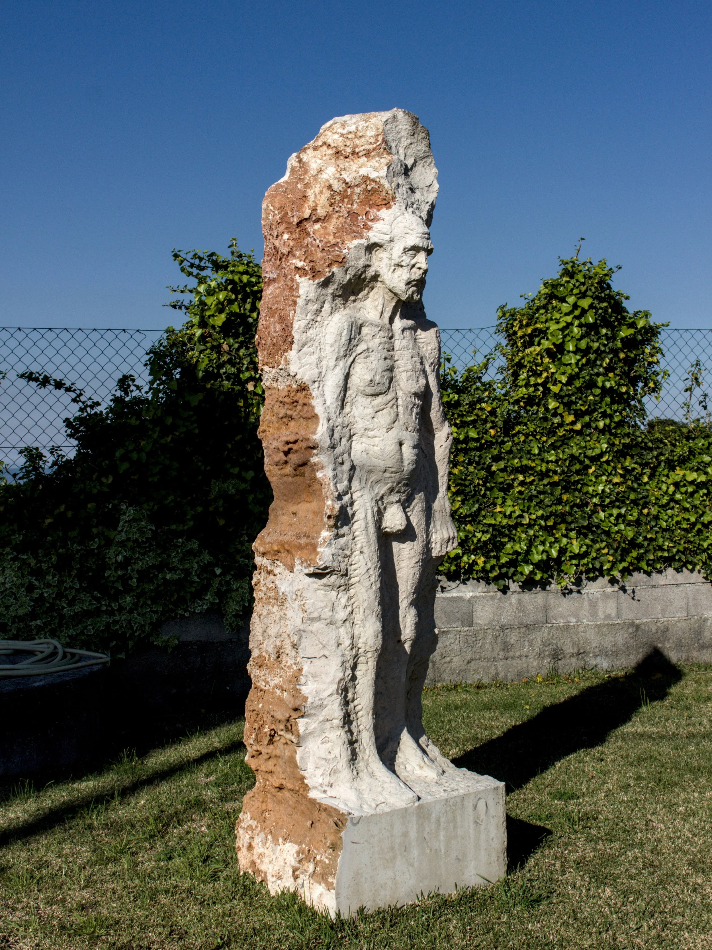 Escultura figurativa de um homem que parece estar com uma expressão séria, feita de pedra, localizada ao ar livre.