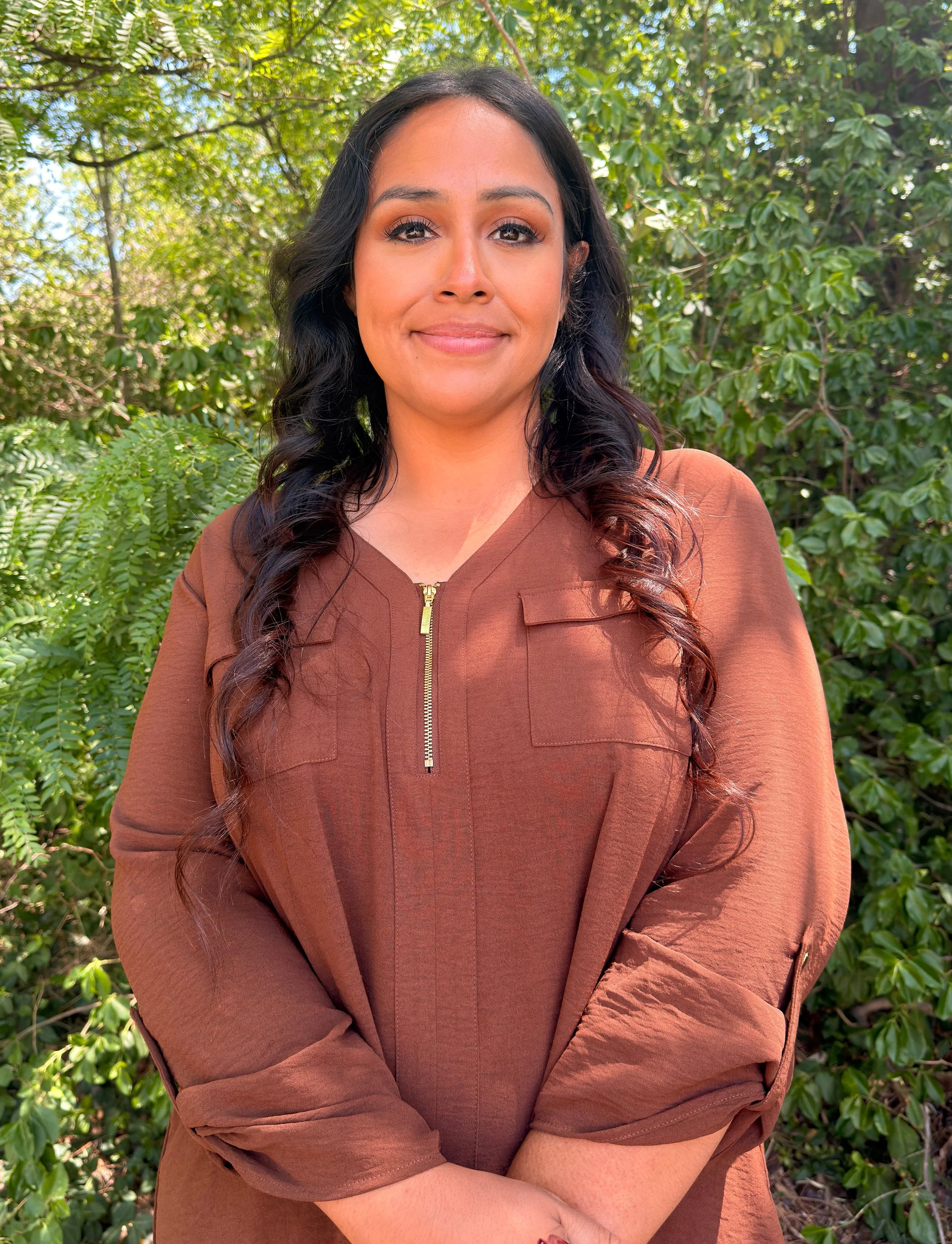 A woman with long dark curly hair wearing a beige turtleneck sweater standing in front of a brick wall.