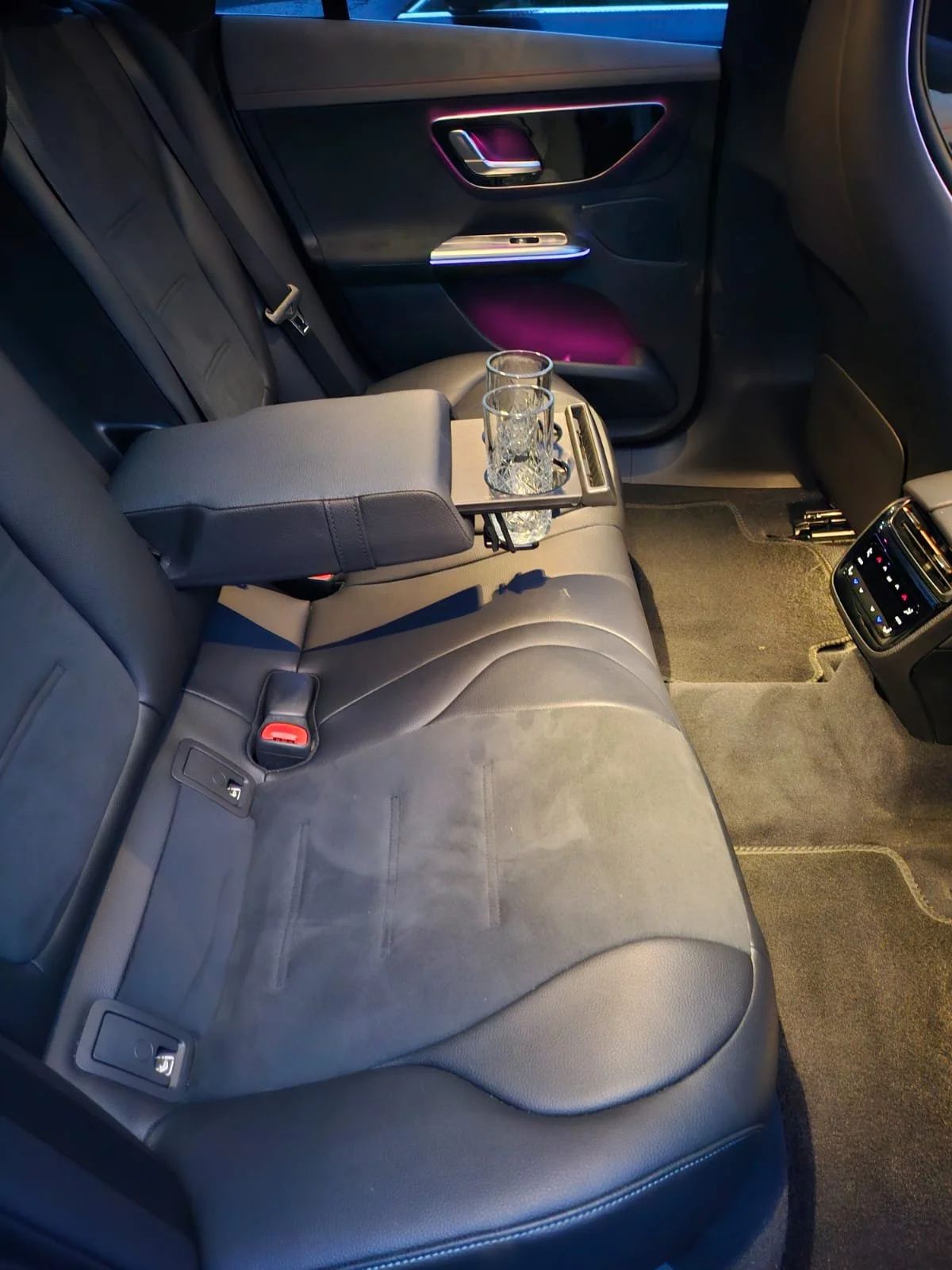 Backseat of a car with two glasses of water on a fold-out tray, black leather seats, and climate control vents on the back of the center console.