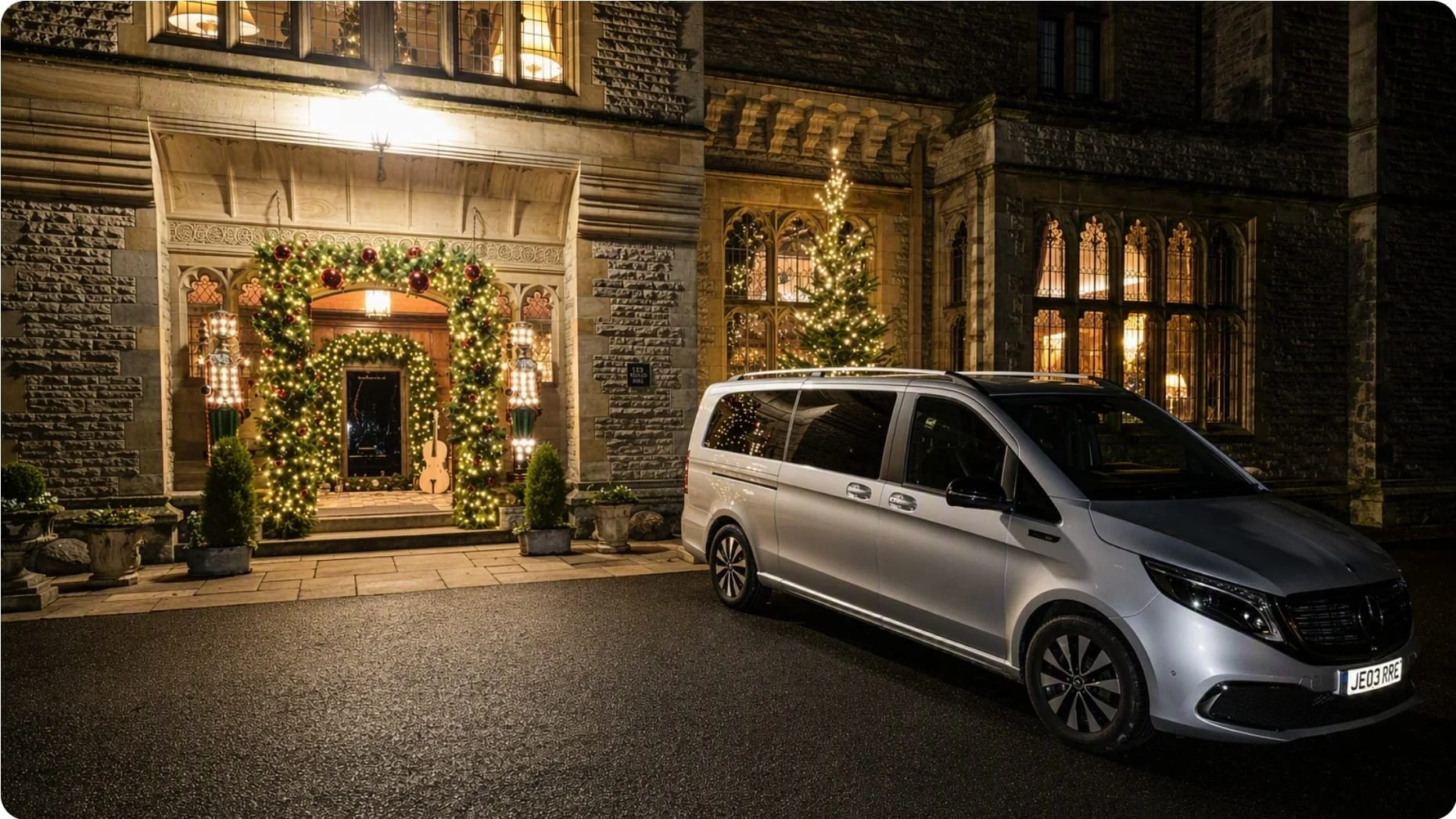 A hotel decorated with holiday lights and Christmas garlands at night, with a Christmas tree visible through the window. A silver Mercedes-Benz EQV is parked in front of Armathwaite Hall Hotel and Spa, Cumbria Lake District