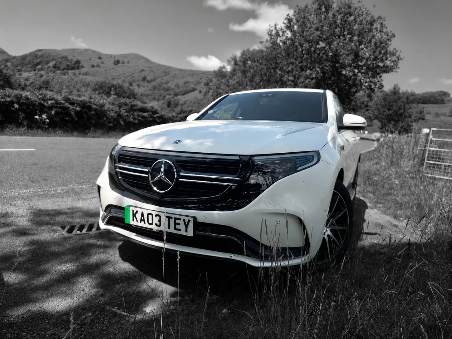 A white Mercedes-Benz car parked on the side of a country road with mountains and trees in the background, black and white filter applied to the scene.