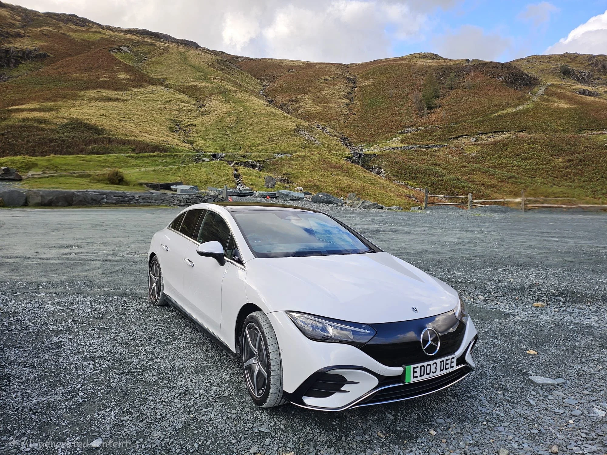 A white Mercedes-Benz car with license plate 'ED03 DEE' parked on a gravel lot in front of a hillside with grassy terrain, rocks, and a cloudy sky.
