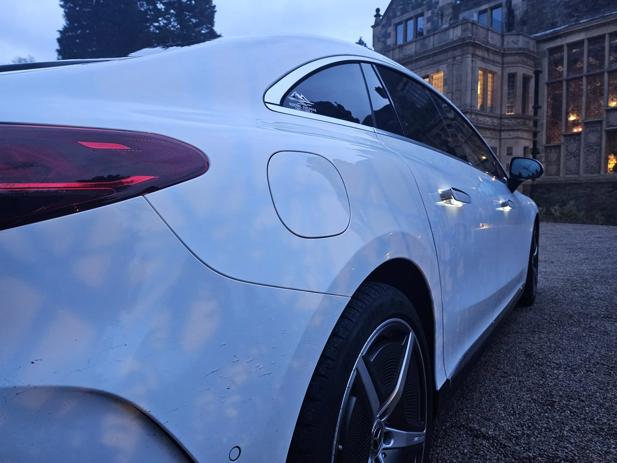 A white luxury car parked outdoors, with a sticker on the side window that reads 'Electric Executive Chauffeur.' In the background, there is a large, historic stone building with illuminated windows, and tall trees are visible against a dusky sky.