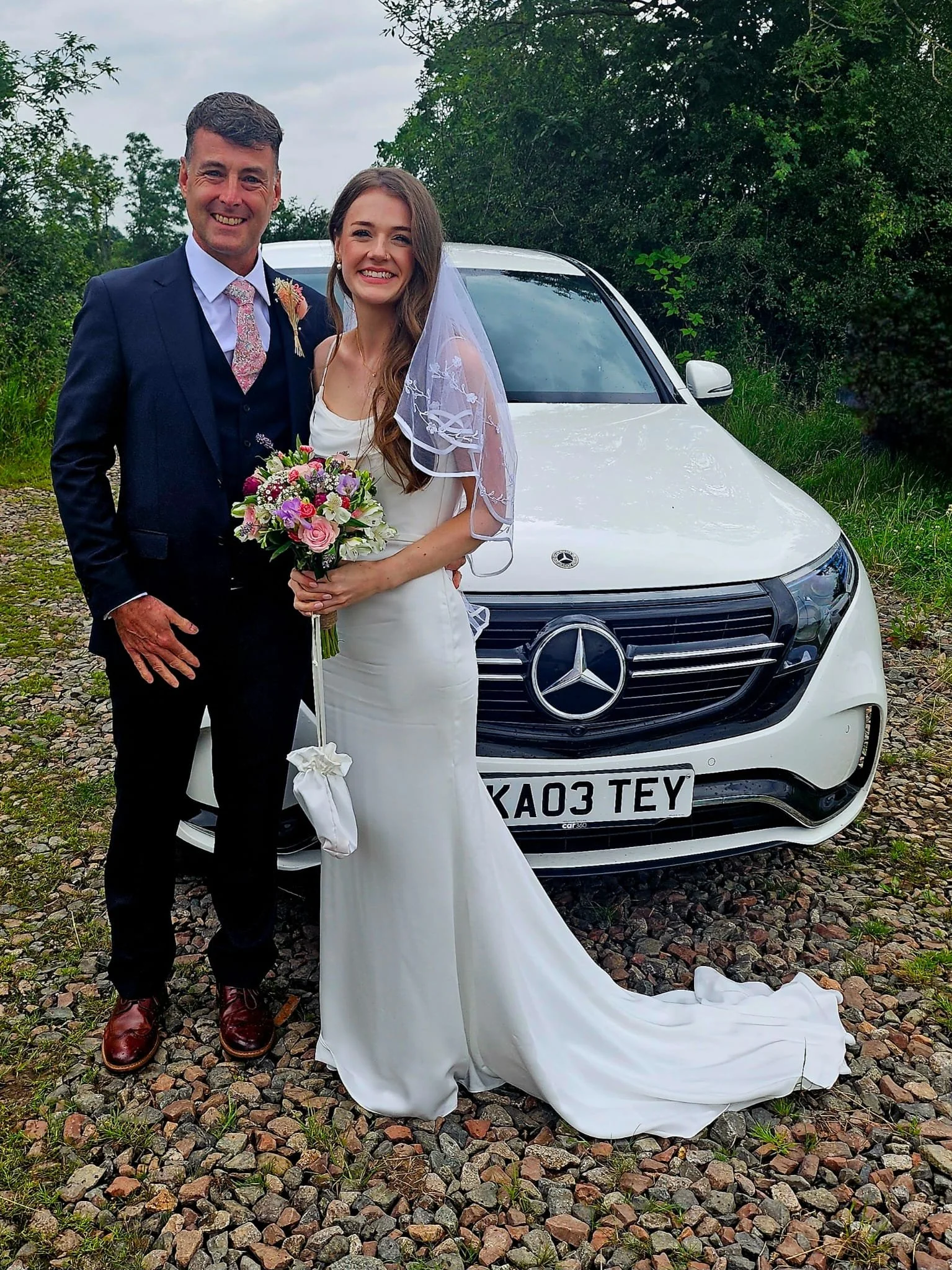 A smiling bride and groom stand in front of a white Mercedes-Benz car. The groom is dressed in a navy suit with a floral tie, and the bride wears a white wedding dress with lace sleeves, holding a bouquet of pink and purple flowers. They are outdoors on a gravel path surrounded by greenery.