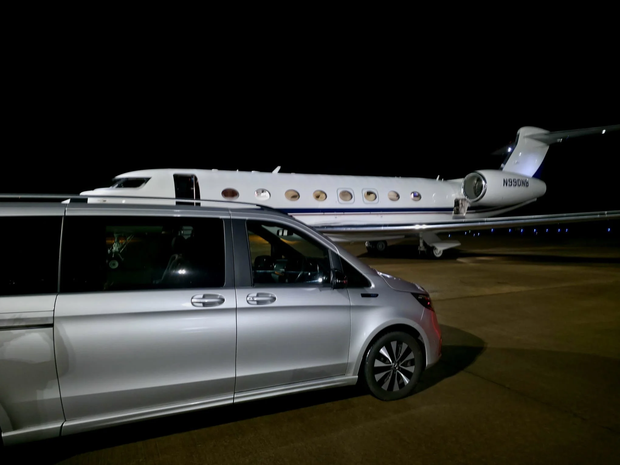 A silver Mercedes-Benz EQV in the foreground and a white private jet at Newcastle Airport in the background at night. Collecting a corporate client.