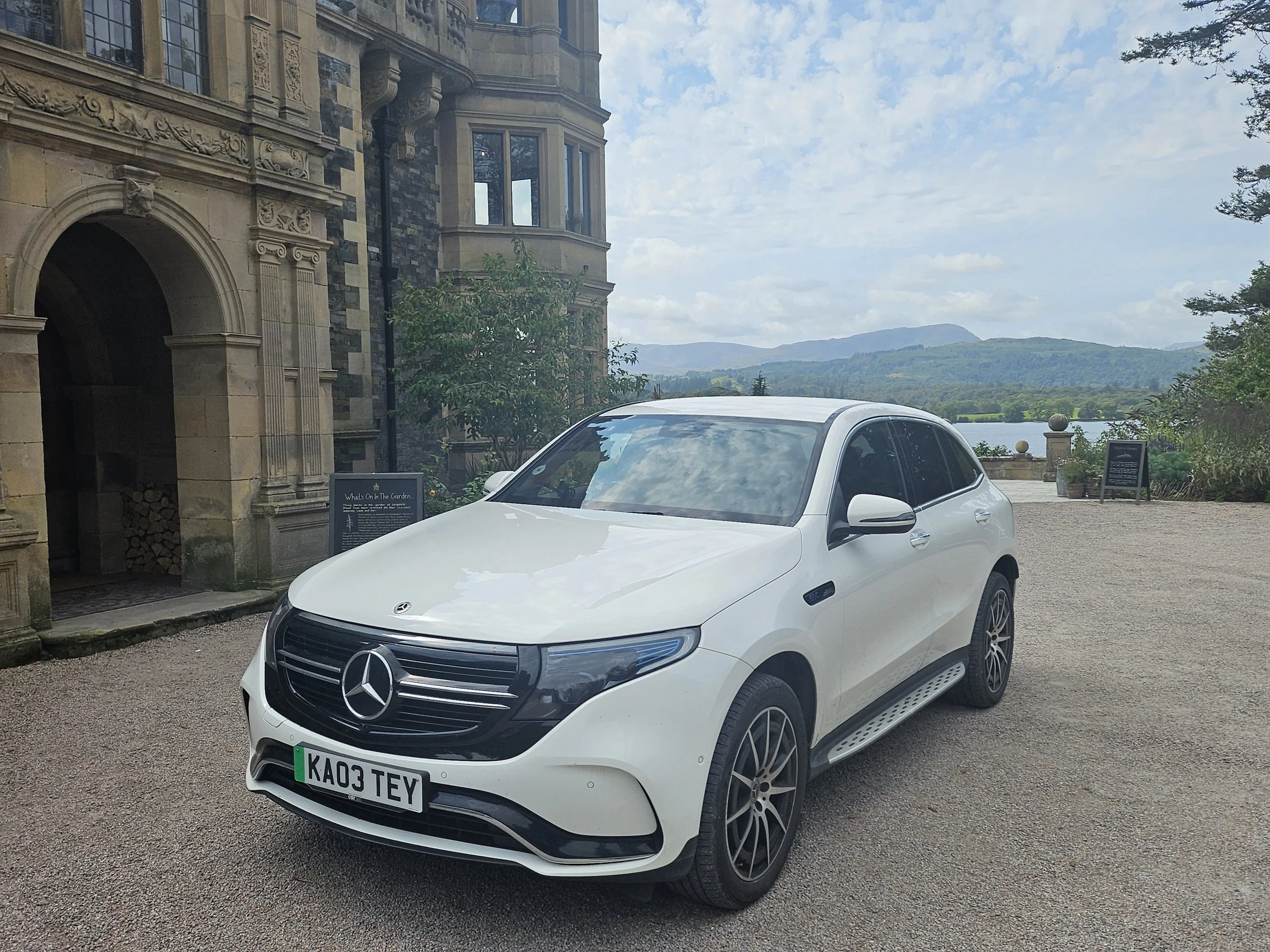 A white Mercedes-Benz car parked outdoors with a scenic background including a historic building with ornate stonework, a sign, trees, a lake, and mountains under a partly cloudy sky.