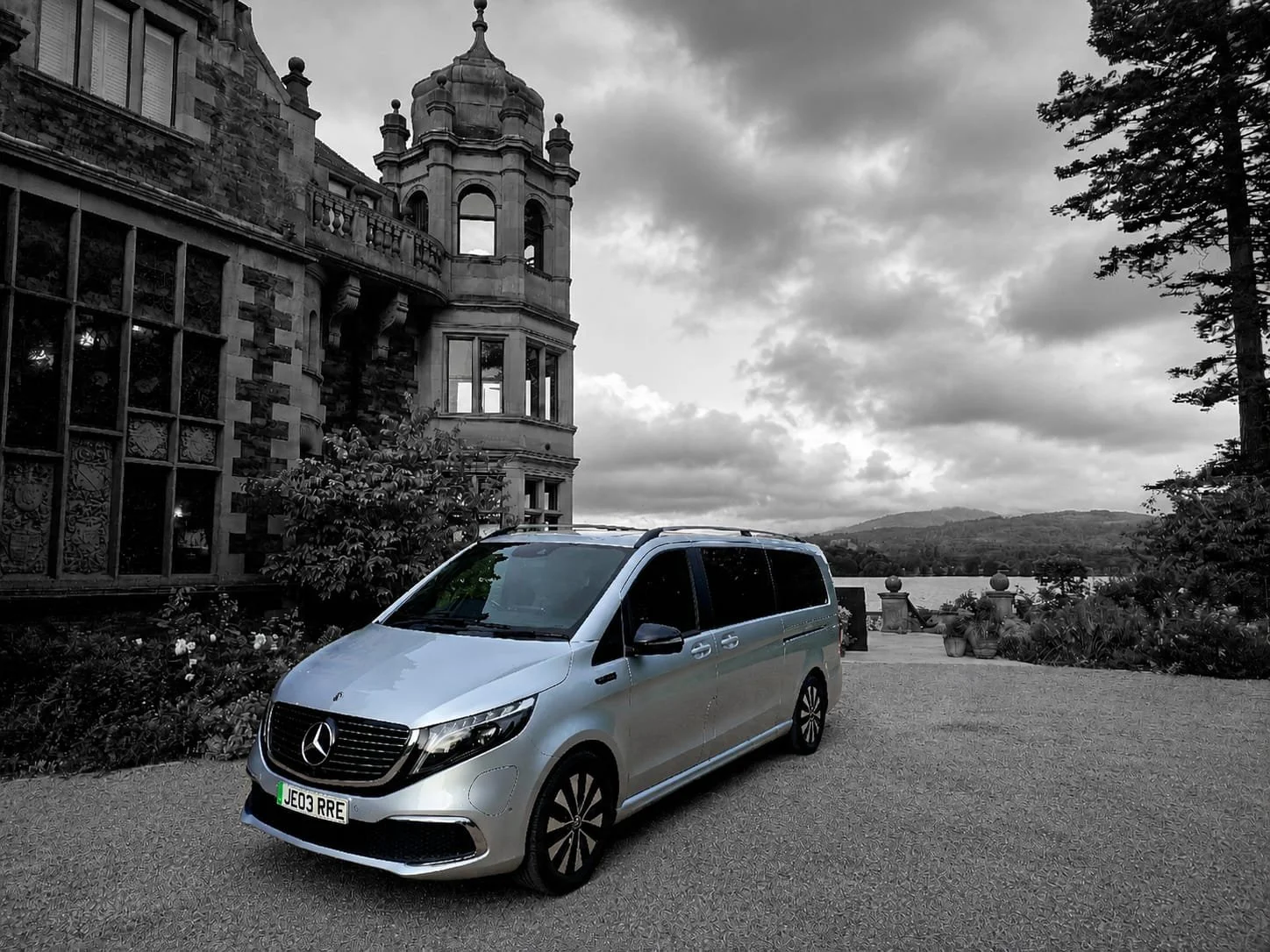 A silver Mercedes-Benz EQV parked on a gravel driveway in front of a historic stone building Langdale Chase Hotel with castle-like architecture, with a lake and hills visible in the background under cloudy skies. Cumbria Lake District