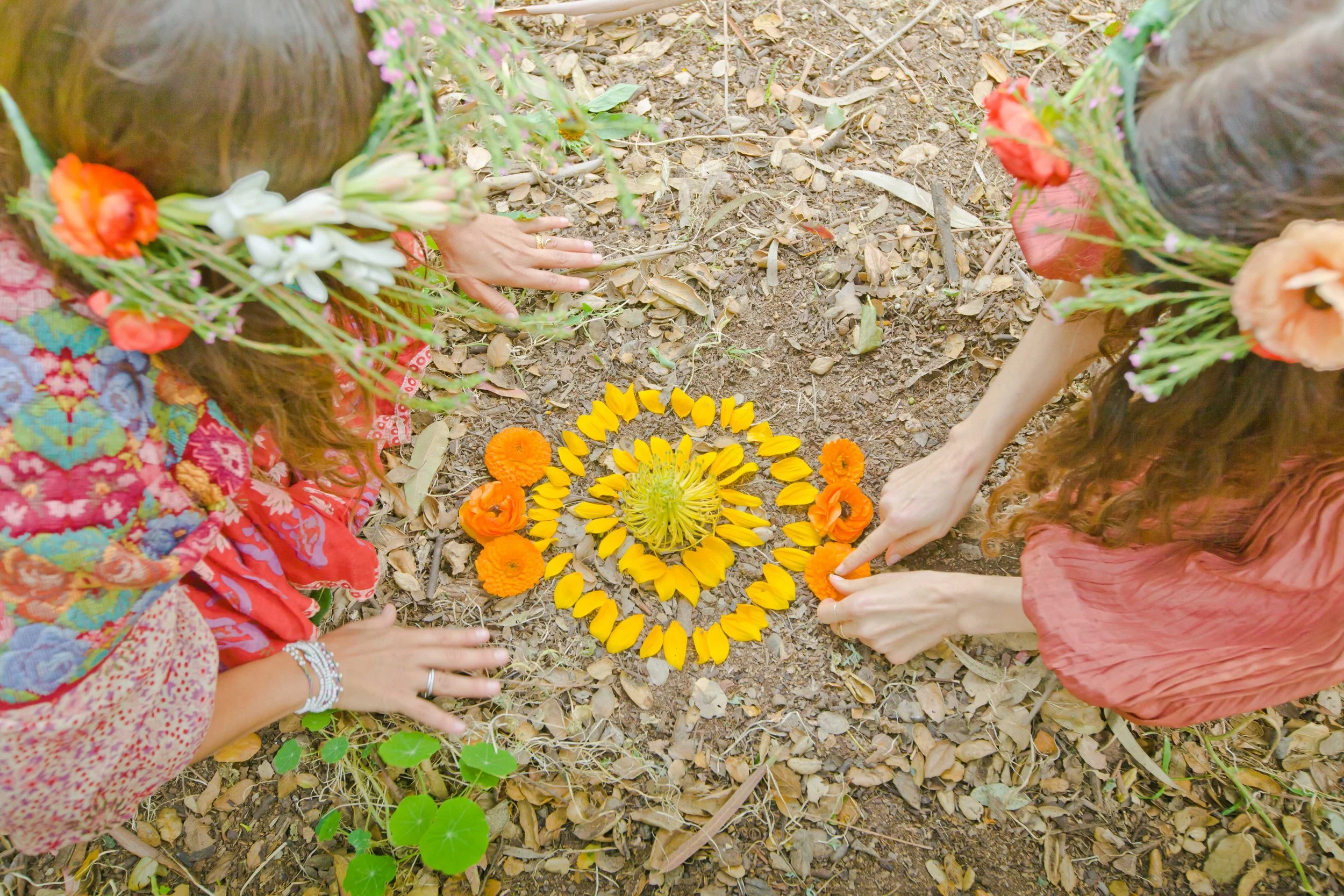Three women with floral headbands and colorful clothing gather on a dirt ground to form a circle with yellow and orange flowers.
