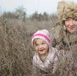 A young girl and an adult, possibly her mother, smiling and playing in a field of tall, dry grass, with the girl wearing a pink and white striped hat.