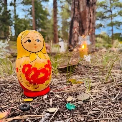 Colorful floral Russian nesting doll standing on a pine-needle forest floor with scattered buttons and trees in the background