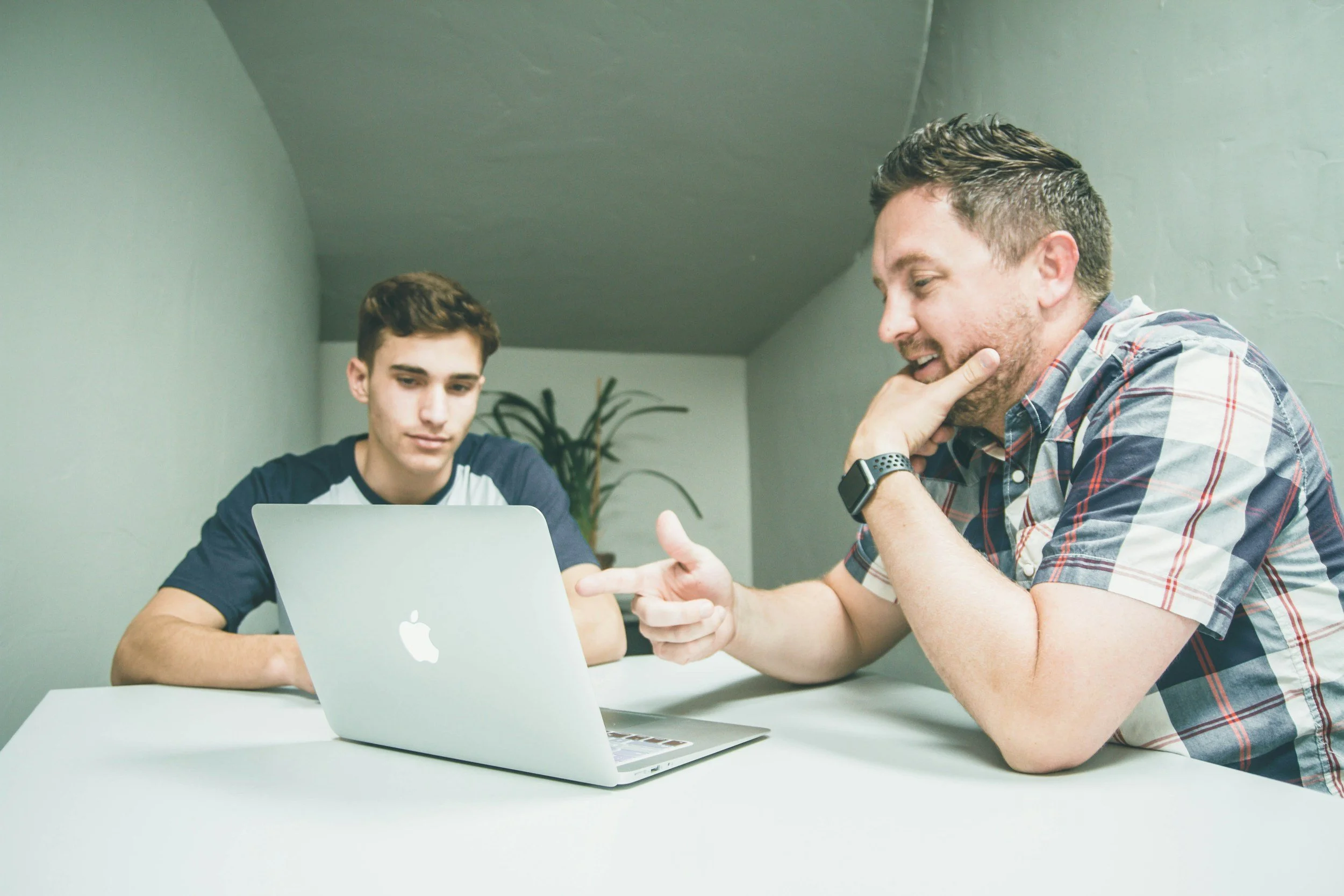 Two men sitting at a table with an open laptop, having a discussion.