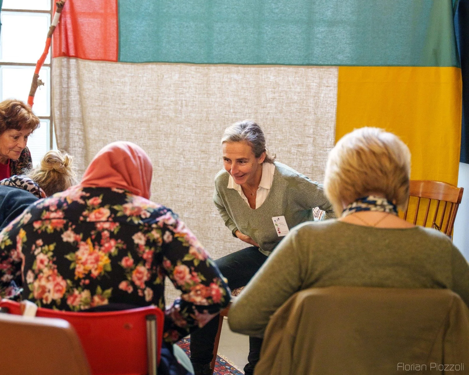 Une femme souriante, en train de parler à un groupe de femmes assises autour d'une table lors d'une réunion ou d'un atelier, avec un fond coloré et un tapis au sol.