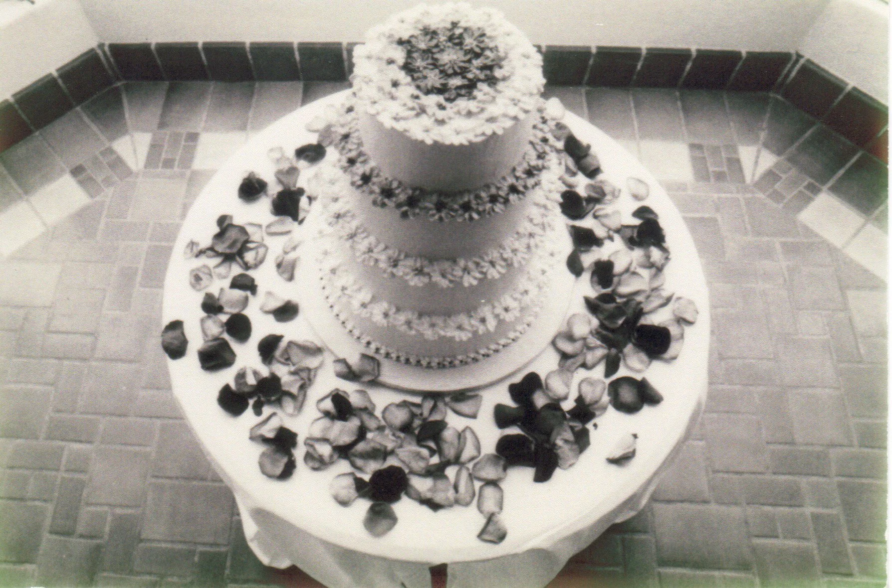 A three-tiered wedding cake with white frosting and floral decorations on each layer, surrounded by scattered rose petals on a round white table.