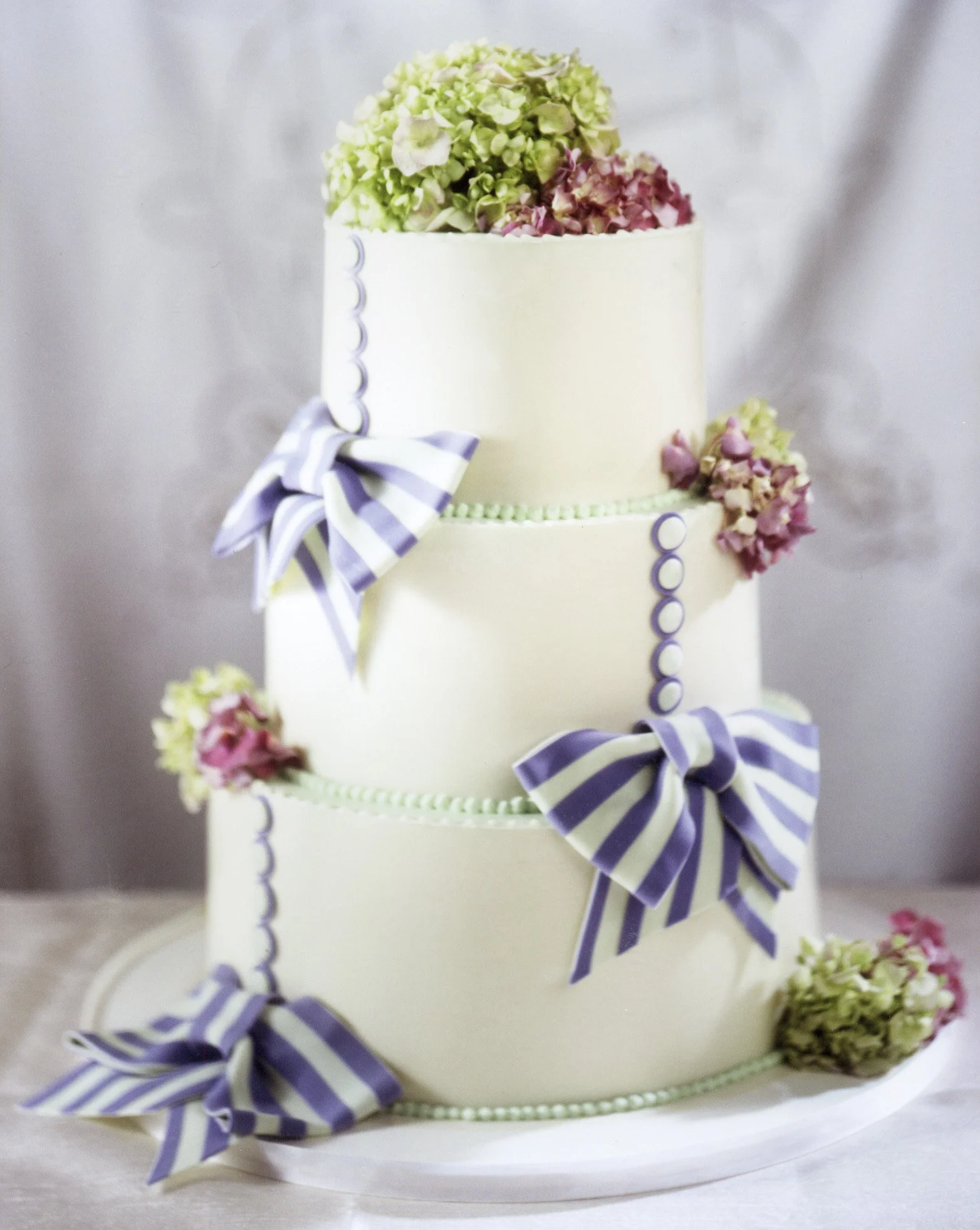 A three-tiered white wedding cake decorated with purple and white striped bows, purple circle embellishments, and pink and green hydrangea flowers.