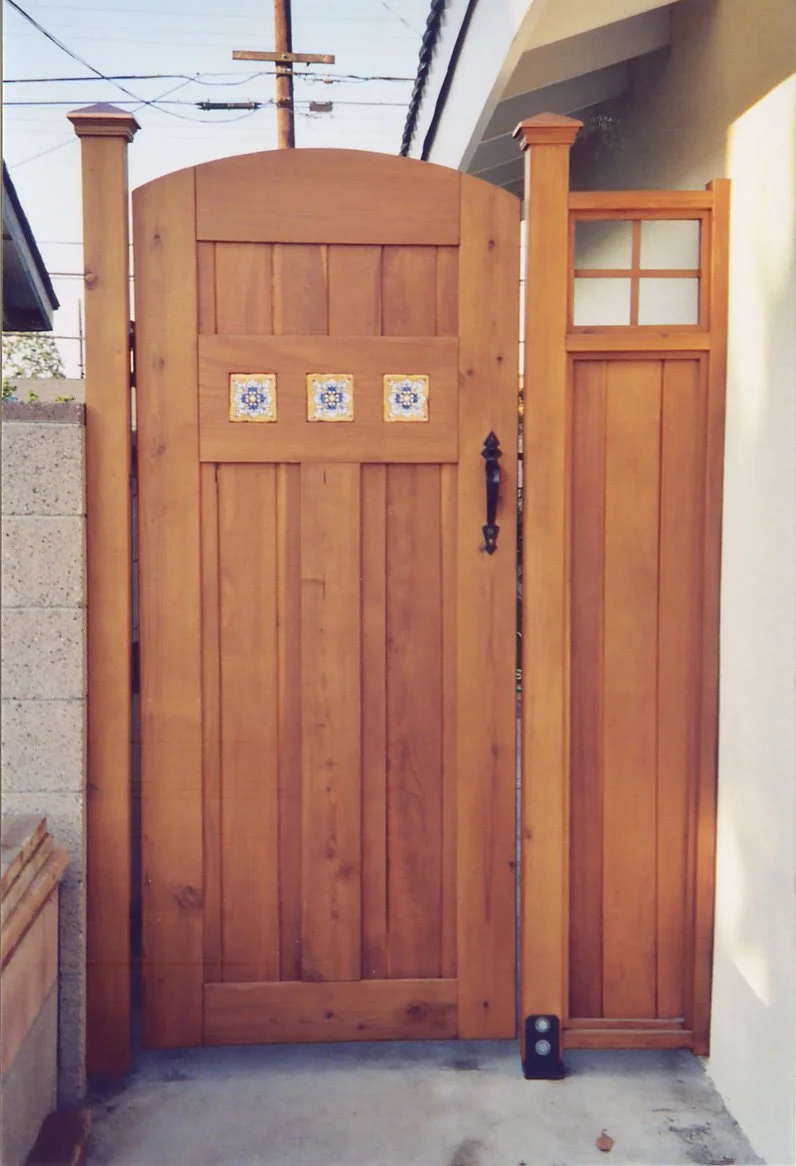 Wooden gate with decorative tiles and a matching wood panel on the right, set in a concrete and brick wall, with a utility pole and power lines visible in the background.