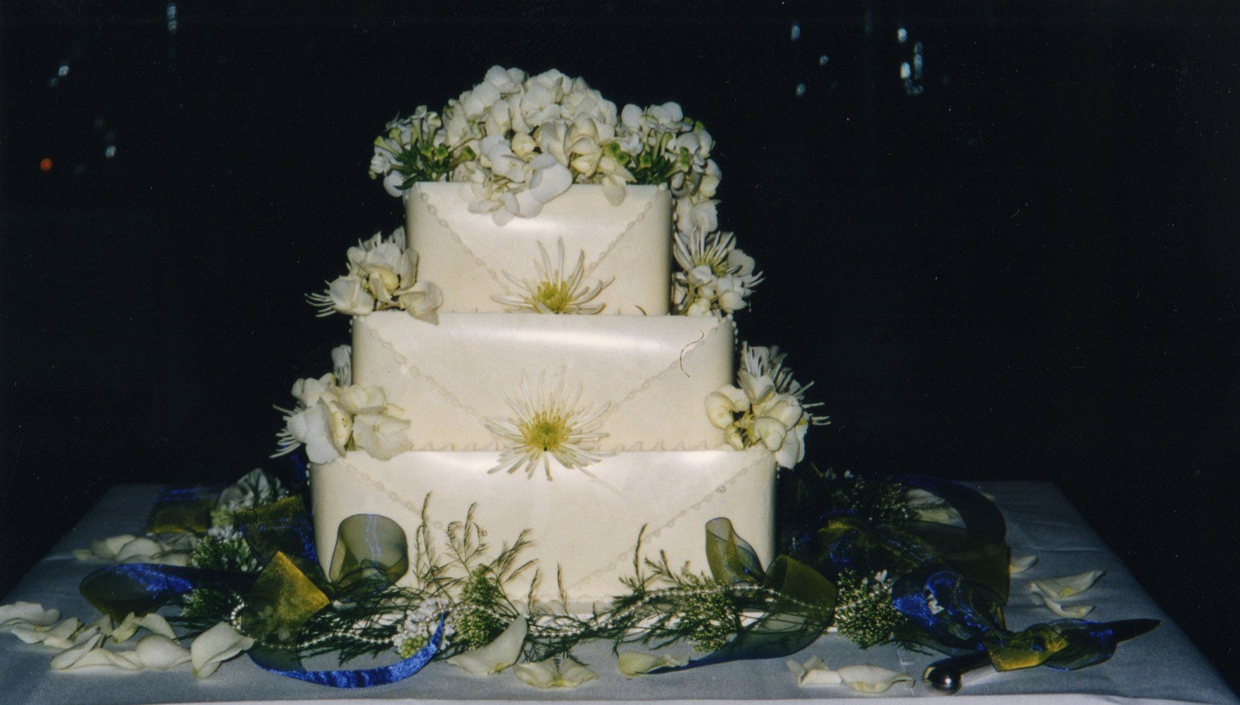 Three-tiered white wedding cake decorated with white flowers and floral patterns, surrounded by flower petals and greenery on a table with dark background.