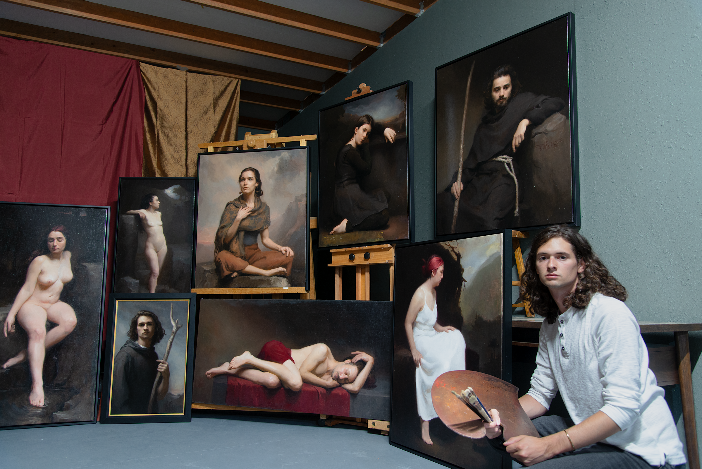 A young male artist with long curly hair sitting on the floor holding a palette and brush, surrounded by several classical-style painted portraits of women and men displayed on easels and leaning against the wall in an art studio.