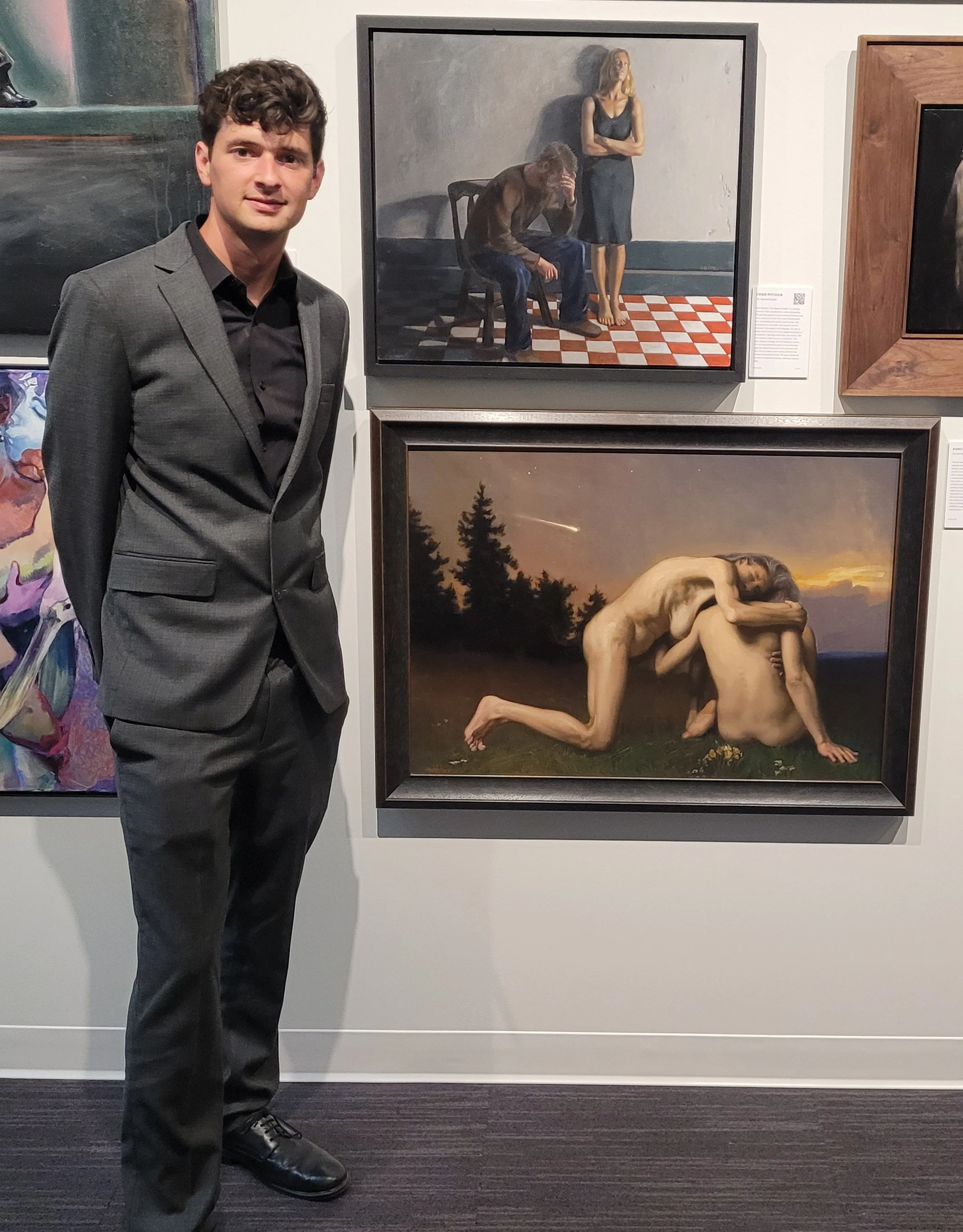 A young man in a gray suit standing in an art gallery, portrait, with paintings on the wall behind him, including one depicting two nude figures outdoors at sunset and another showing a surreal scene with a person with a monkey face and a woman.