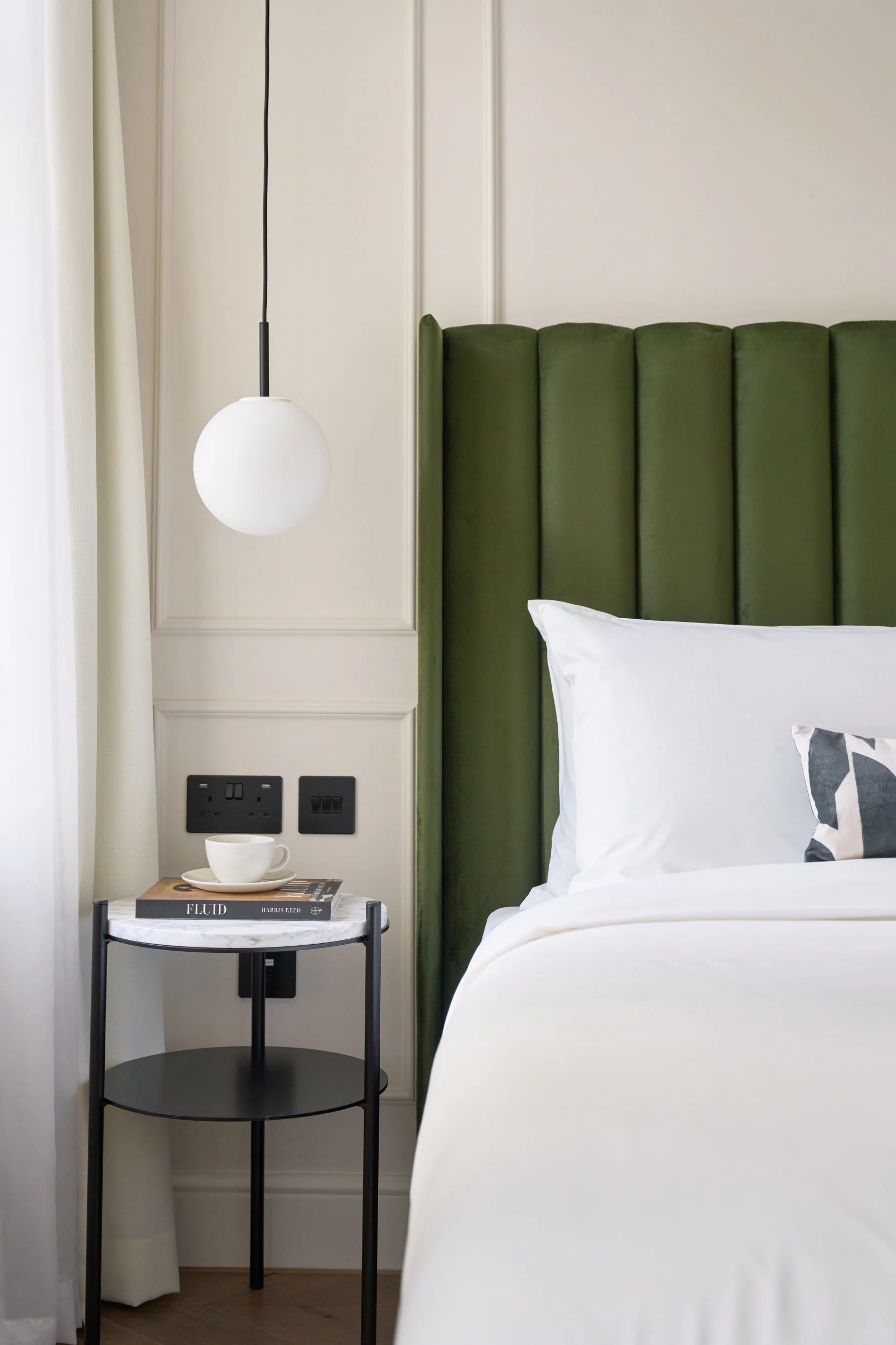 Close-up of a bedroom corner with a green upholstered headboard, white bedding, a small round side table with a marble top holding a cup, saucer, and books, and a wall-mounted spherical white bedside lamp.