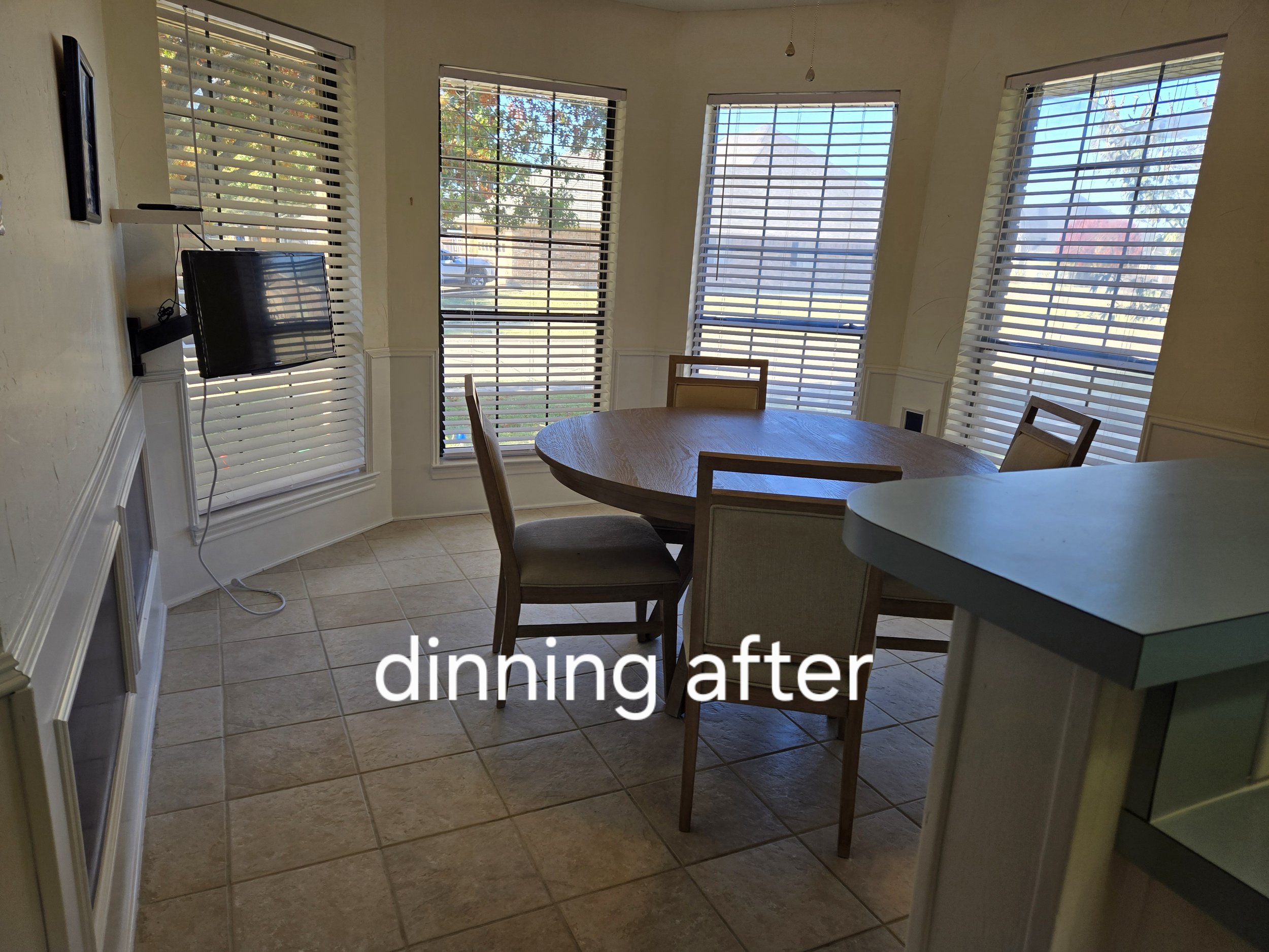 Dining room with round table, chairs, wall-mounted TV, and multiple windows with blinds.