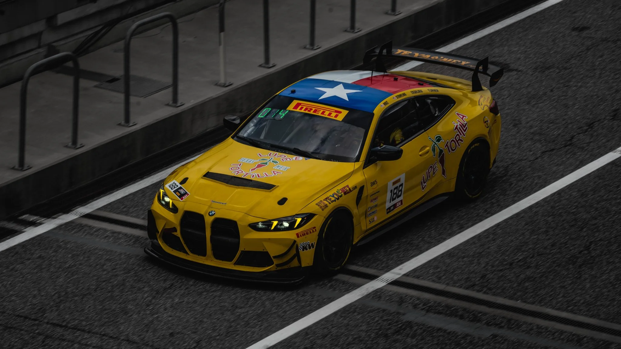 A yellow BMW race car with the Texas flag on the hood and various sponsor stickers, parked in a pit lane on a racetrack.