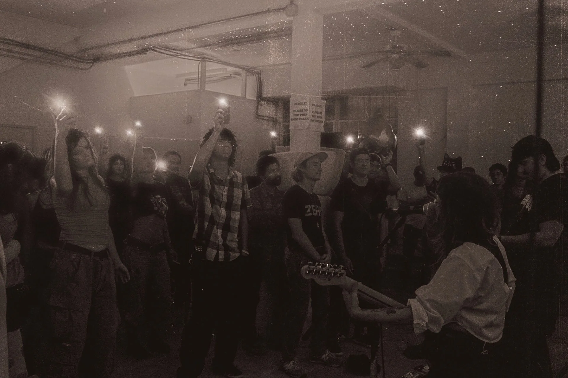 Group of people standing indoors, some holding up their phones with flashlights on, gathered in a crowded space with a person holding a guitar and woman with a hat in the foreground.
