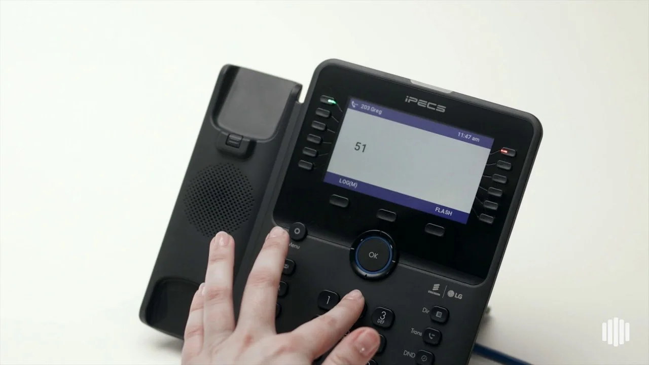A person dialing a number on a black iPECS office phone with a colour LCD screen.