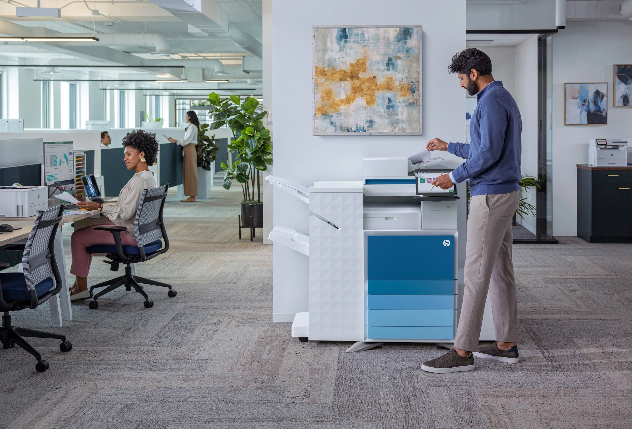 Office worker using an HP multifunction printer in a modern office with multiple employees working at cubicles and a woman walking in the background.