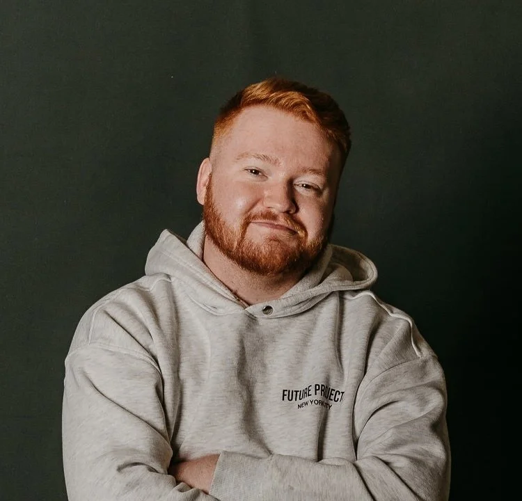 A man with ginger hair and beard, wearing a light gray hoodie with "Future Project New York City" printed on it, smiling with arms crossed against a dark background.