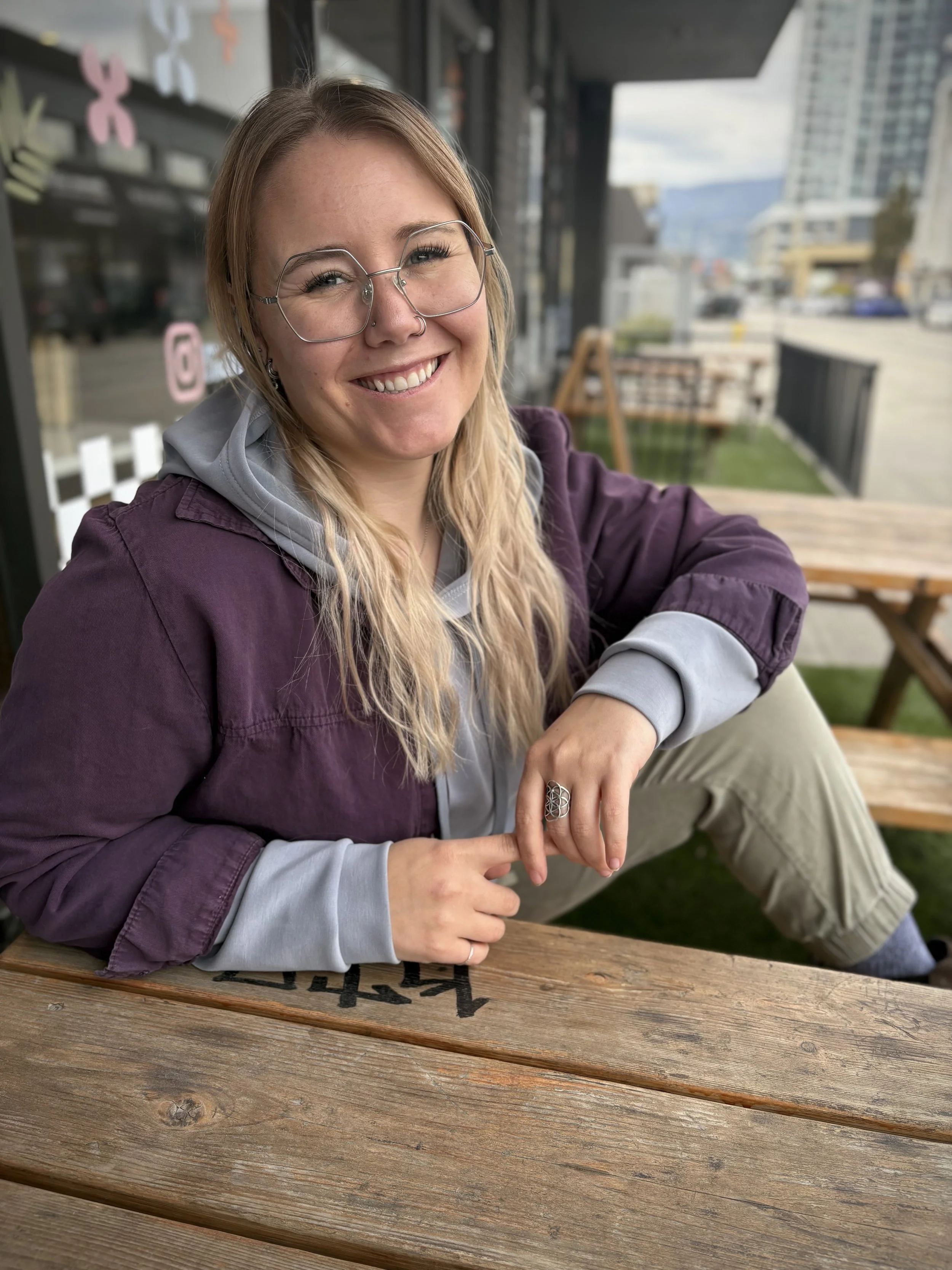 A smiling woman with glasses and long blonde hair sitting outdoors at a wooden table with a city skyline in the background.