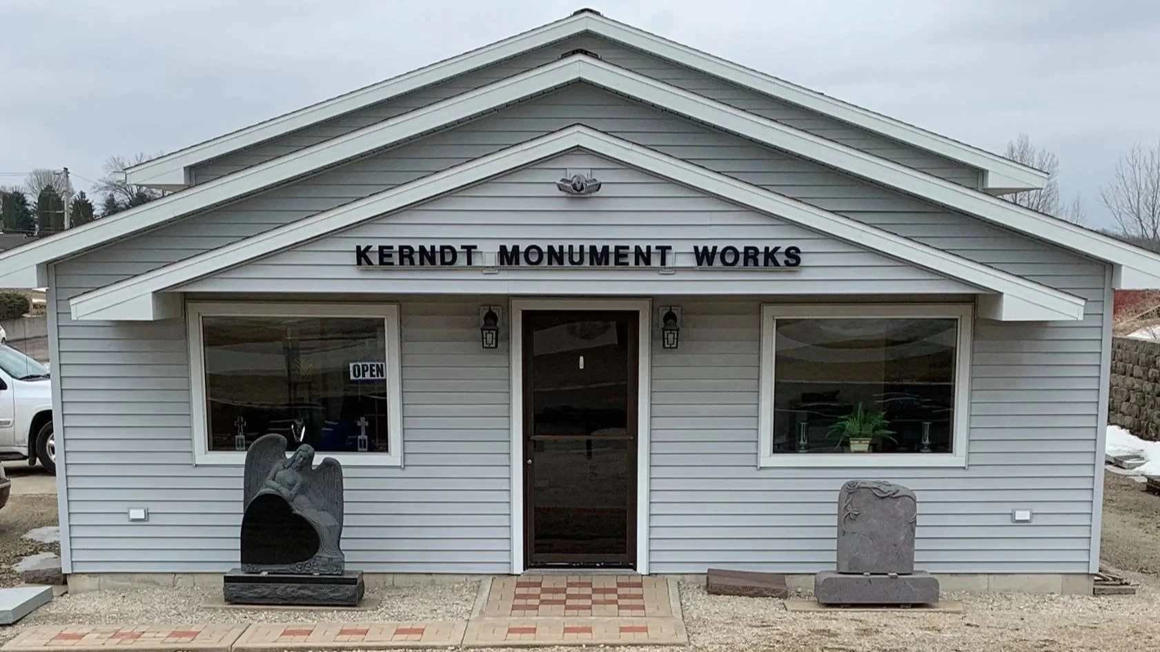 Kerndt Monument Works in Waukon Iowa. Front view of a building labeled 'Kerndt Monument Works' with a glass door, two windows, a stone sculpture of a angel, and a stone memorial plaque outside.