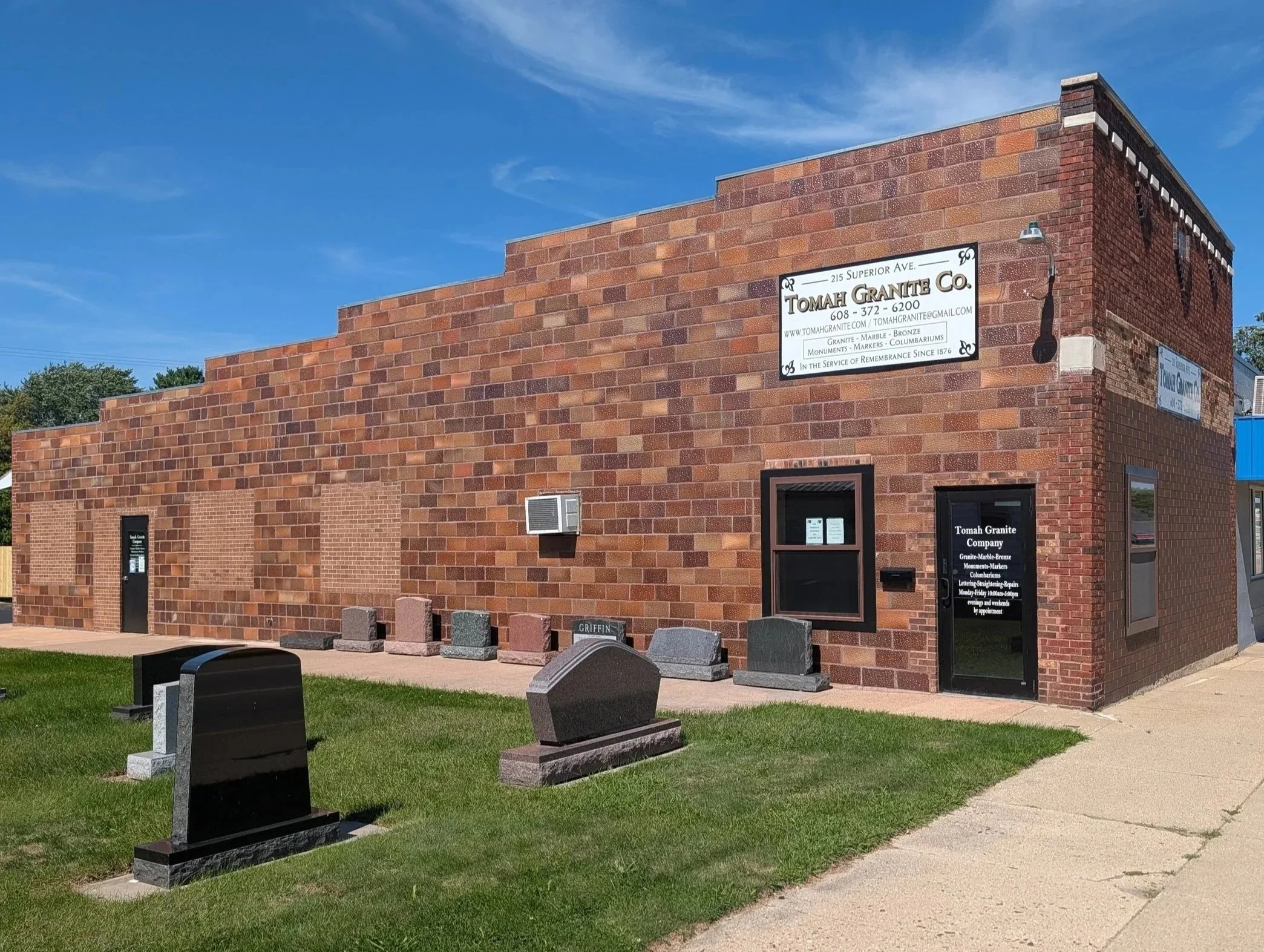 Tomah Granite Company in Tomah Wisconsin. A brick building with small tombstones in front and a sidewalk on the right side. The building has a small window, an air conditioning unit, and a sign for the Tomah Granite Compnay.