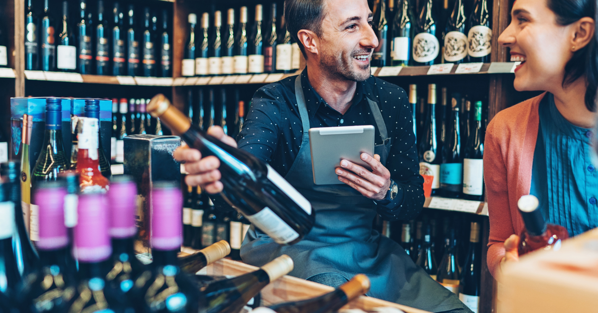 A man and woman shopping for wine in a liquor store, smiling and talking, with shelves of wine bottles behind them.