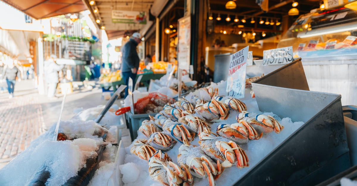 Seafood display at an outdoor market with oysters on ice, a sign for whole cooked Dungeness crab, and other seafood items, with a person shopping in the background.