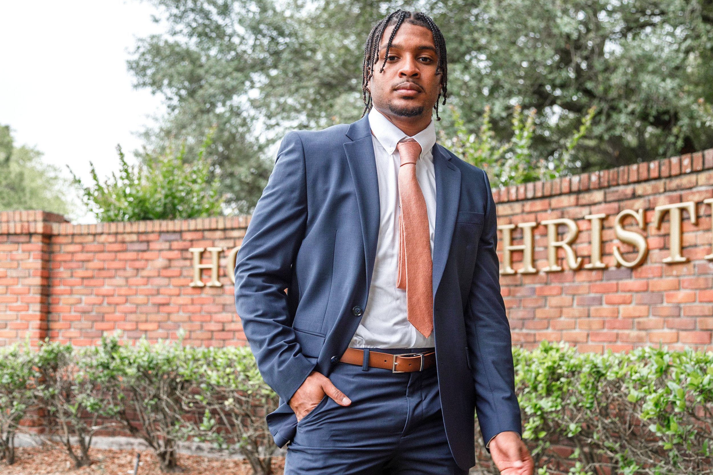 A man in a navy suit and light brown tie standing outdoors in front of a brick wall with the partial sign 'H*RES*'. He has his right hand in his pocket and a confident expression, with trees and greenery in the background.