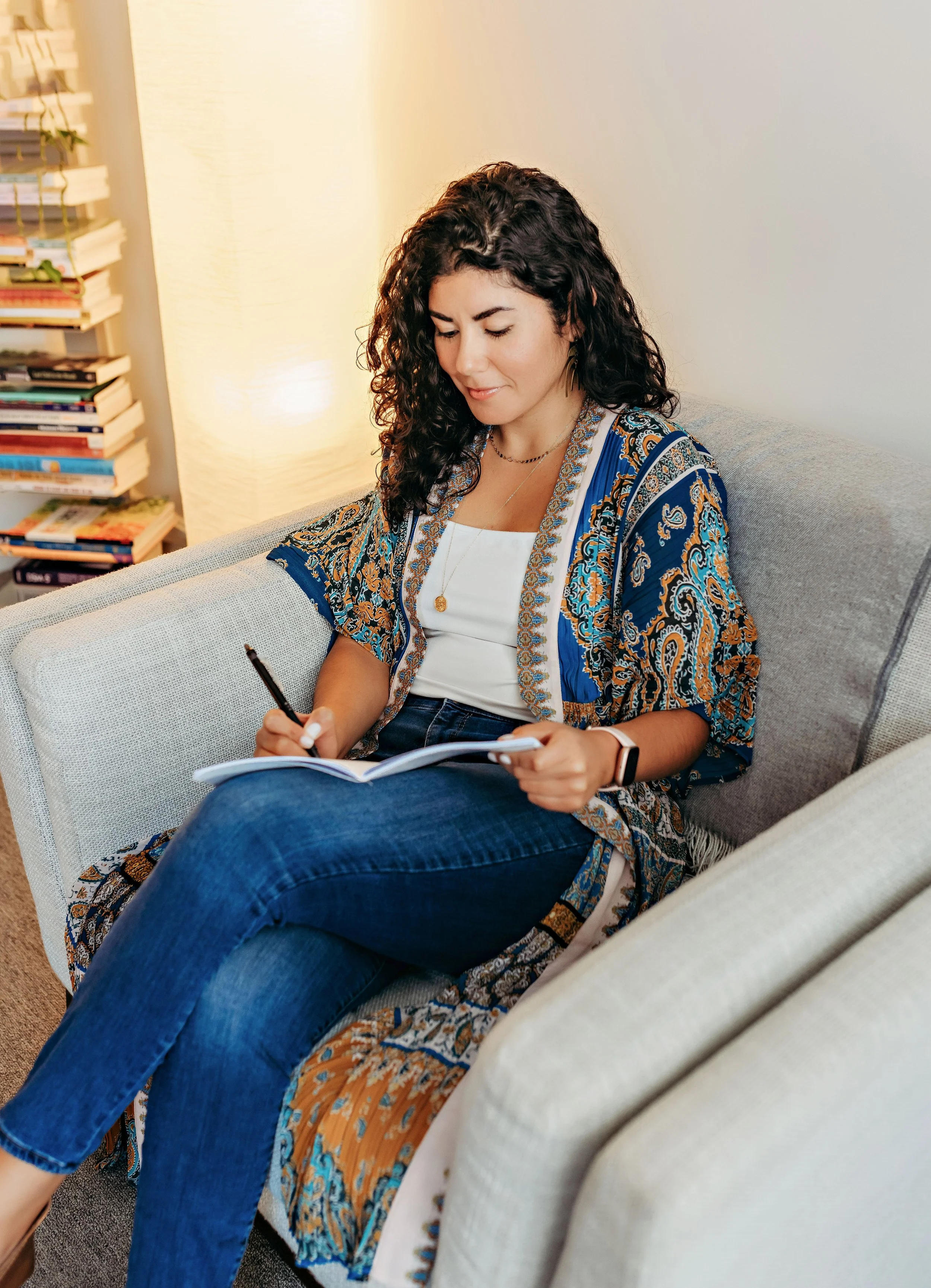 Young woman sitting on a beige sofa with a patterned shawl, writing in a notebook with a pen, near a bookcase filled with books.