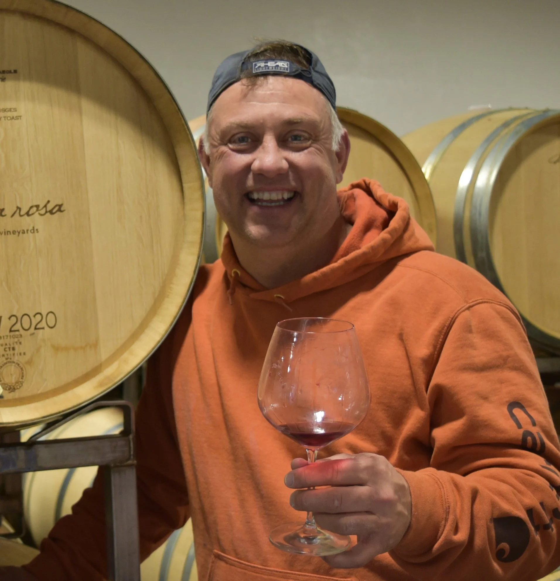 Alma Rosa assistant winemaker, Tony Biagi, wearing a backwards hats, holding a wine glass with red wine in it, standing in front of a rack of wine barrels.