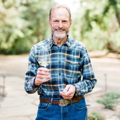 Image of Alma Rosa founder, Richard Sanford, holding a glass of chardonnay wine wearing a plaid shirt, blue jeans, and a leather belt with belt buckle.