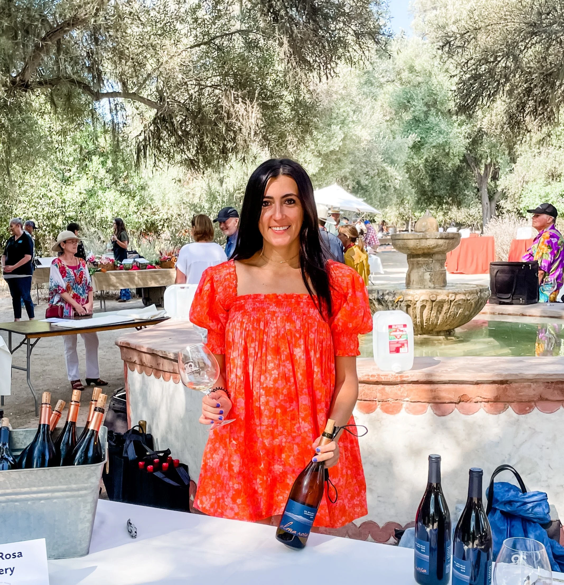 Alma Rosa Winery head winemaker, Samra Morris, wearing a red dress, smiling, holding a wine glass and a wine bottle on a table with a wine tablecloth and other wine bottles on the table.
