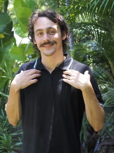 young man in black polo shirt with moustache smiling and tapping under his collarbone with both hands