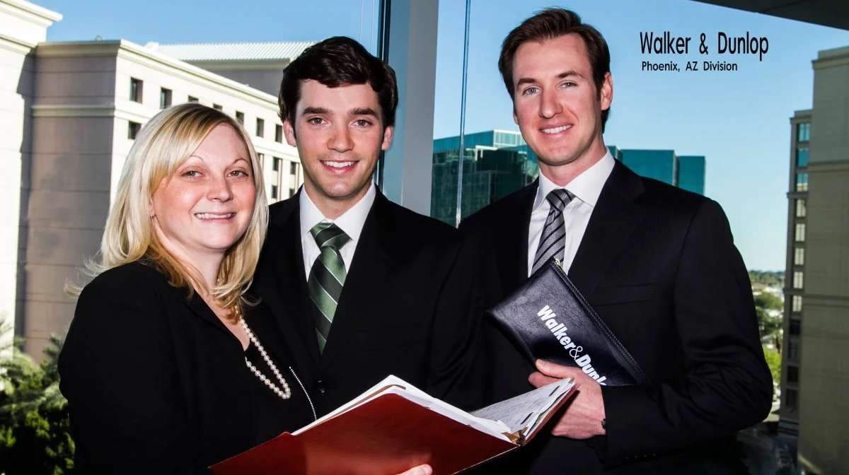 Three professionals standing in front of a window with city buildings in the background. They are dressed in business attire, holding documents. The woman on the left is blonde and smiling, the man in the middle has dark hair with a green striped tie