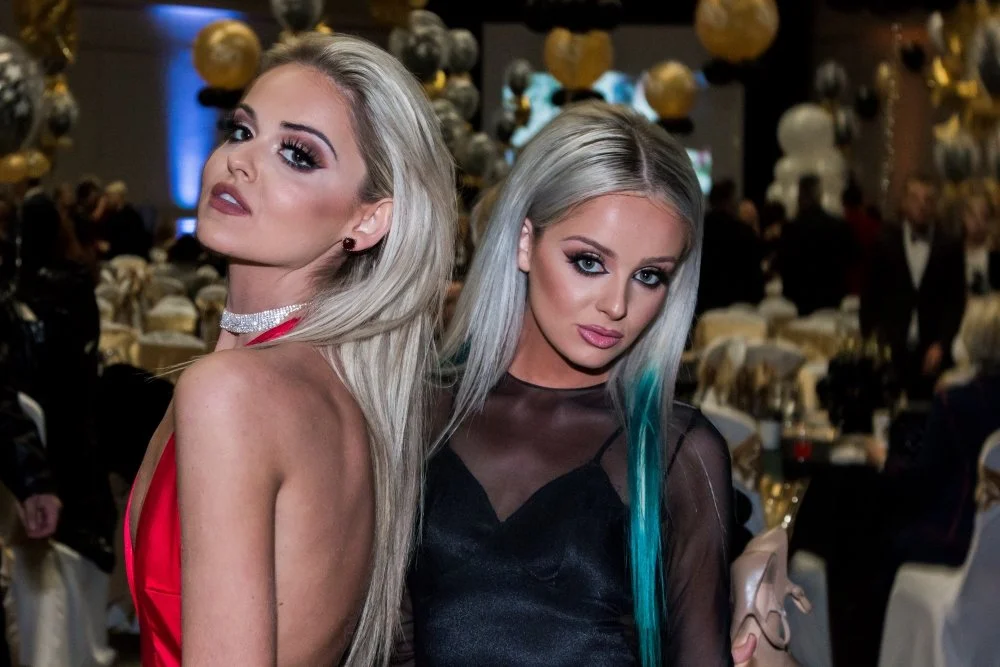 Two glamorous women with platinum blonde hair posing at a formal event with a decorated background and seated guests.