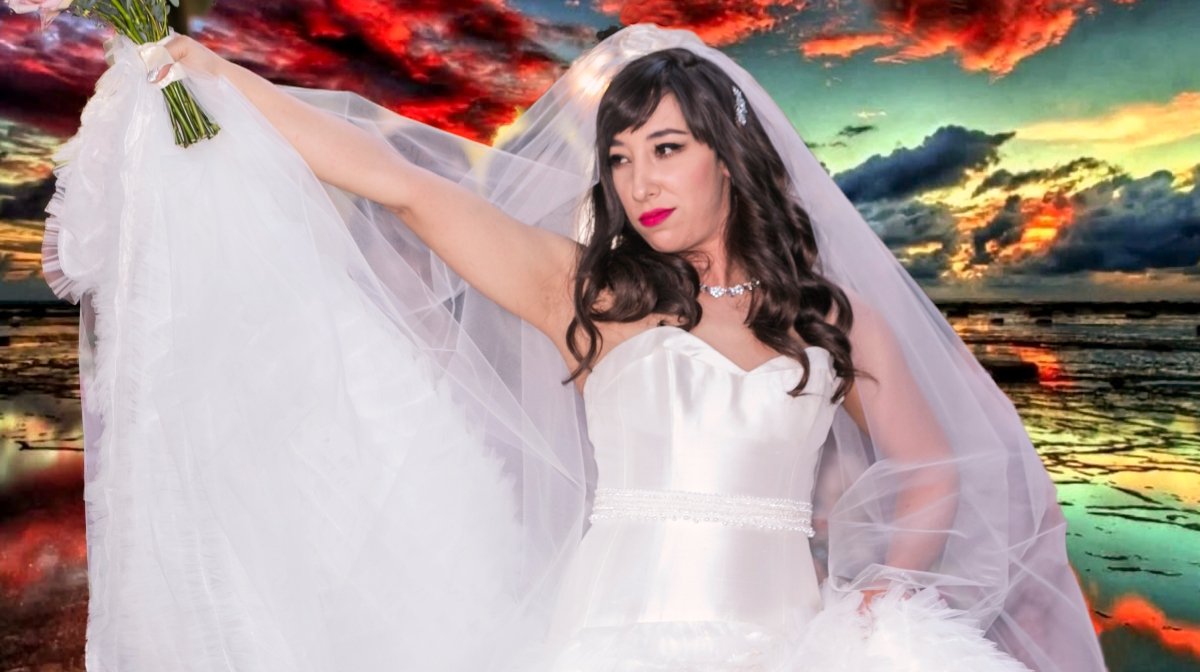 A woman in a white wedding gown and veil holding a bouquet against a colorful sunset over a beach.