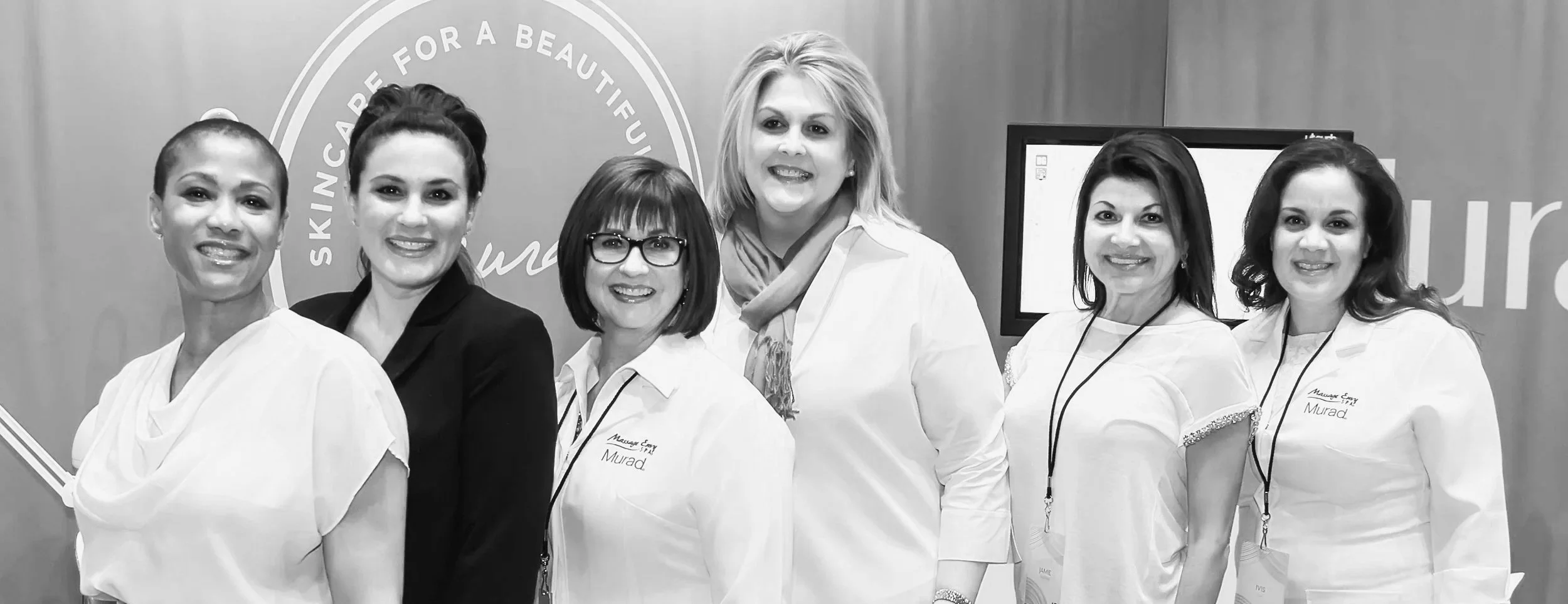 Seven women, some wearing white coats and others in professional attire, standing together in front of a backdrop with partially visible text and logo for a skin care event.