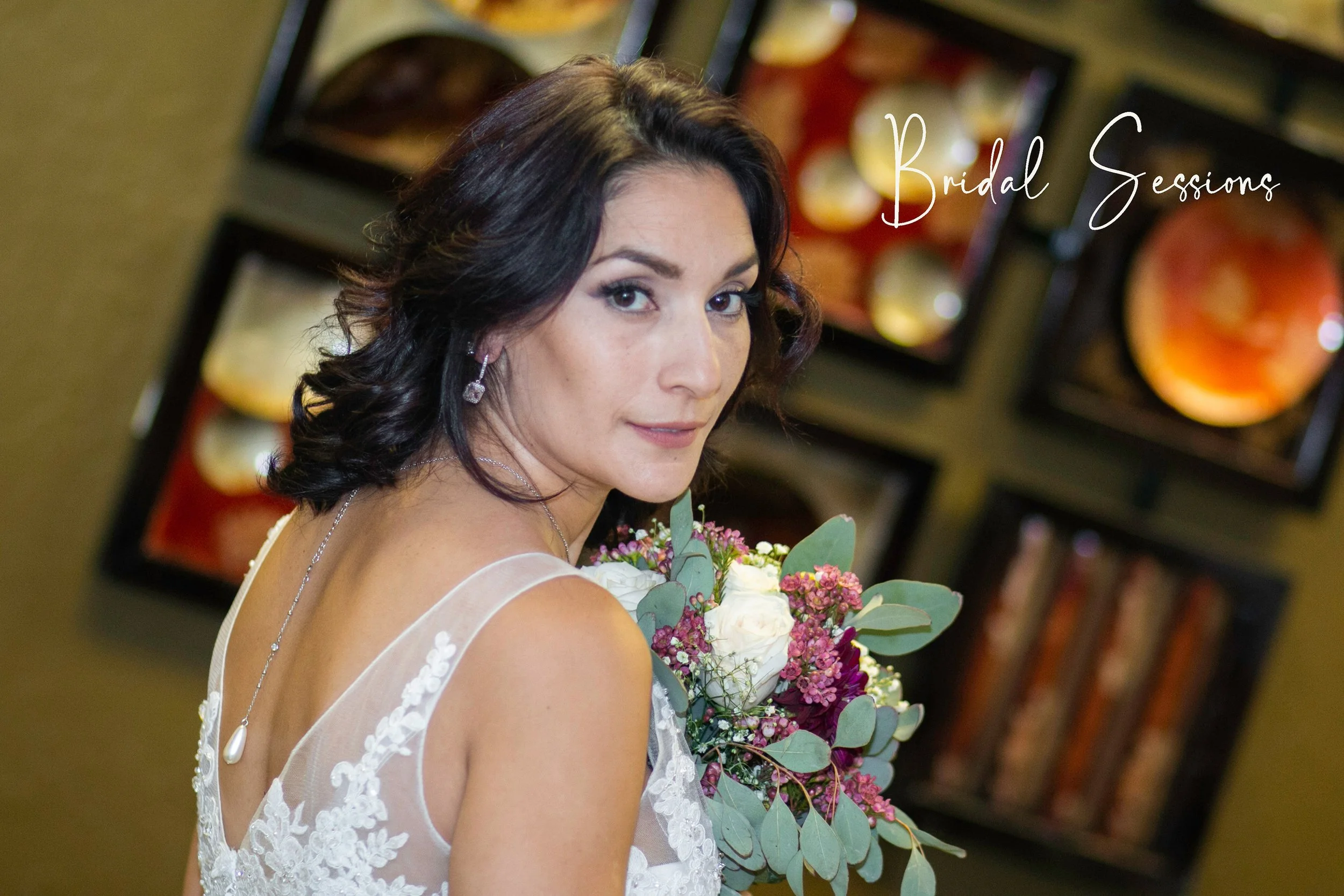 A bride with dark hair holding a bouquet of white and pink flowers, wearing a white lace wedding dress with sheer straps, standing indoors with framed artwork in the background and the words 'Bridal Sessions' written in the upper right corner.