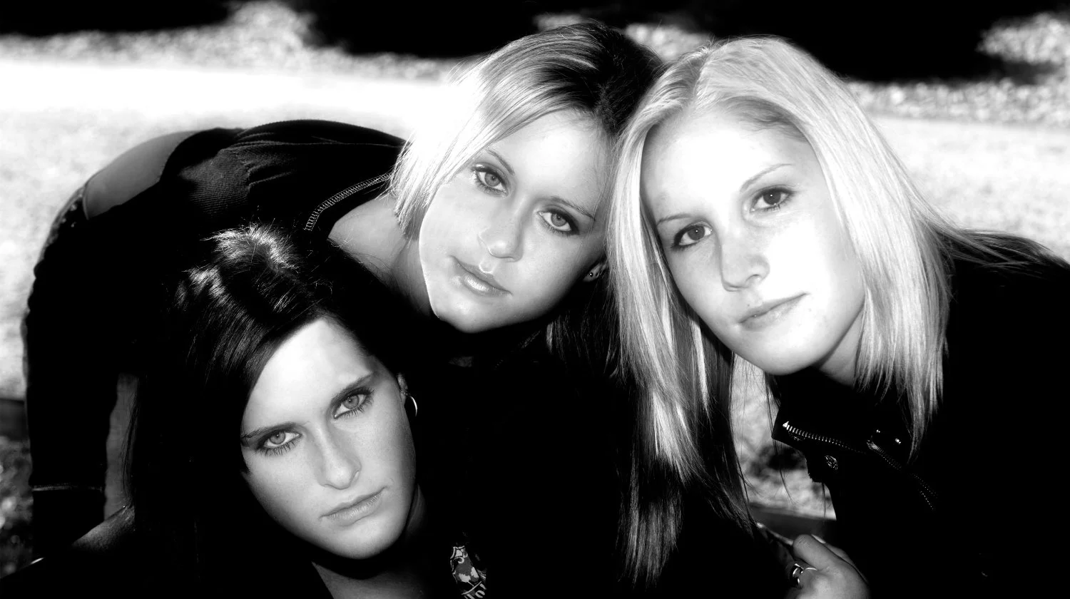 Three young women with blonde and dark hair lying close together outdoors, looking at the camera.